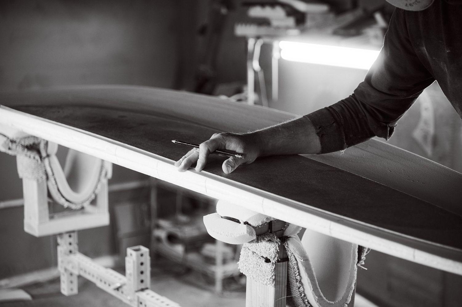 A surfboard shaper carefully runs his hand along the rail of a dark surfboard.