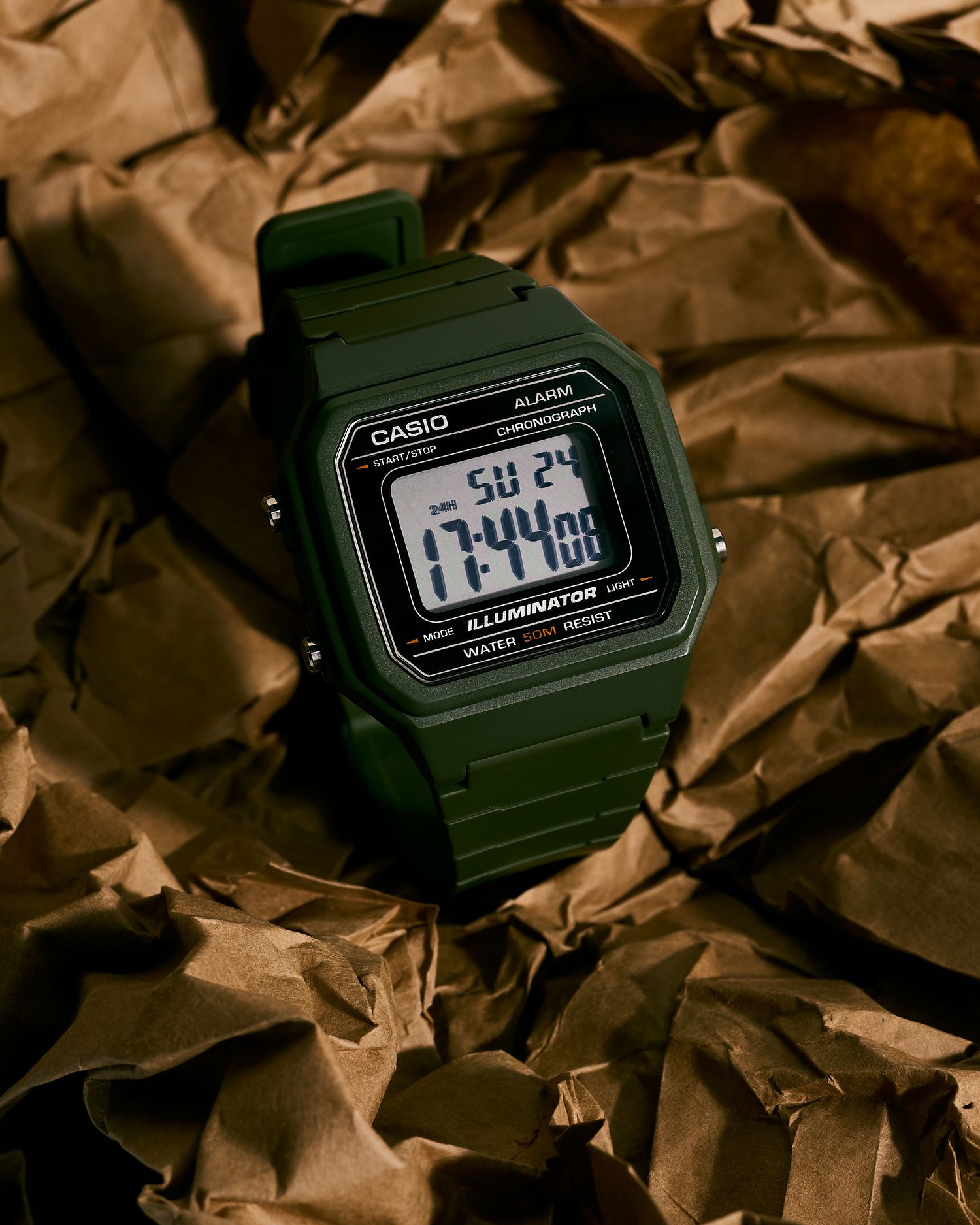 Green Casio digital alarm chronograph watch with illuminator display showing 11:44, placed on crumpled brown paper.