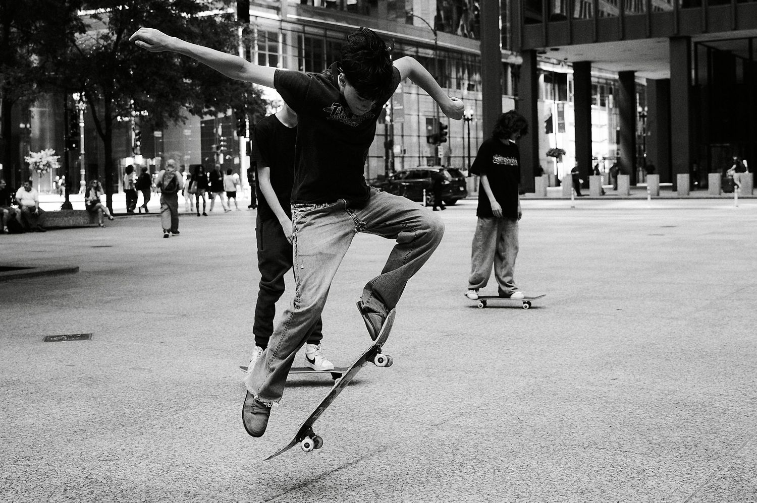 Black and white photo of a skateboarder mid-kickflip in a busy urban plaza with spectators nearby.