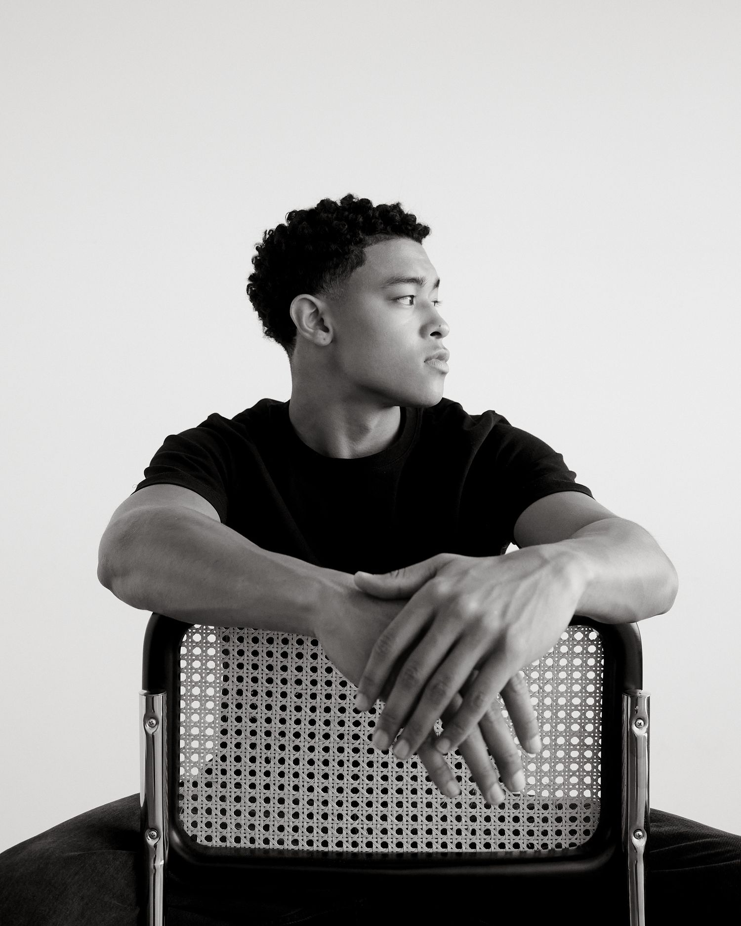Black and white portrait of a young man in a black t-shirt seated backwards on a cane chair.