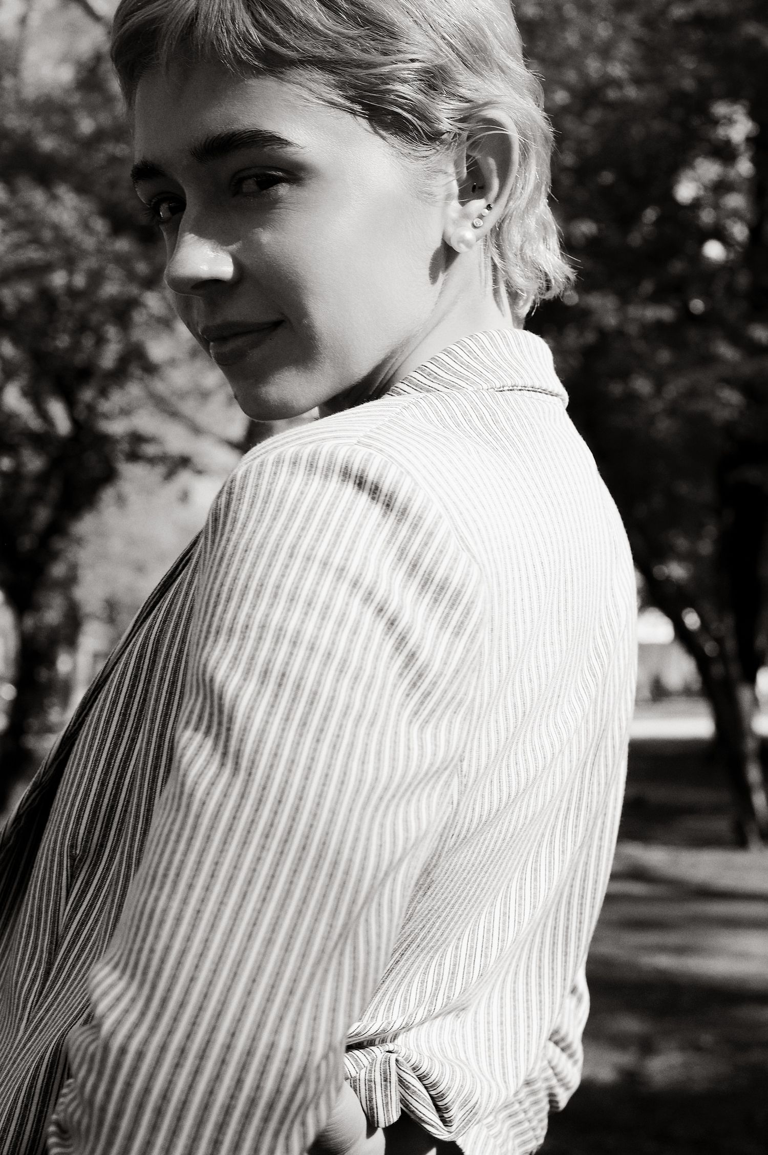 Young blonde woman in pinstripe blazer glances back over shoulder in sunlit park, black and white portrait.