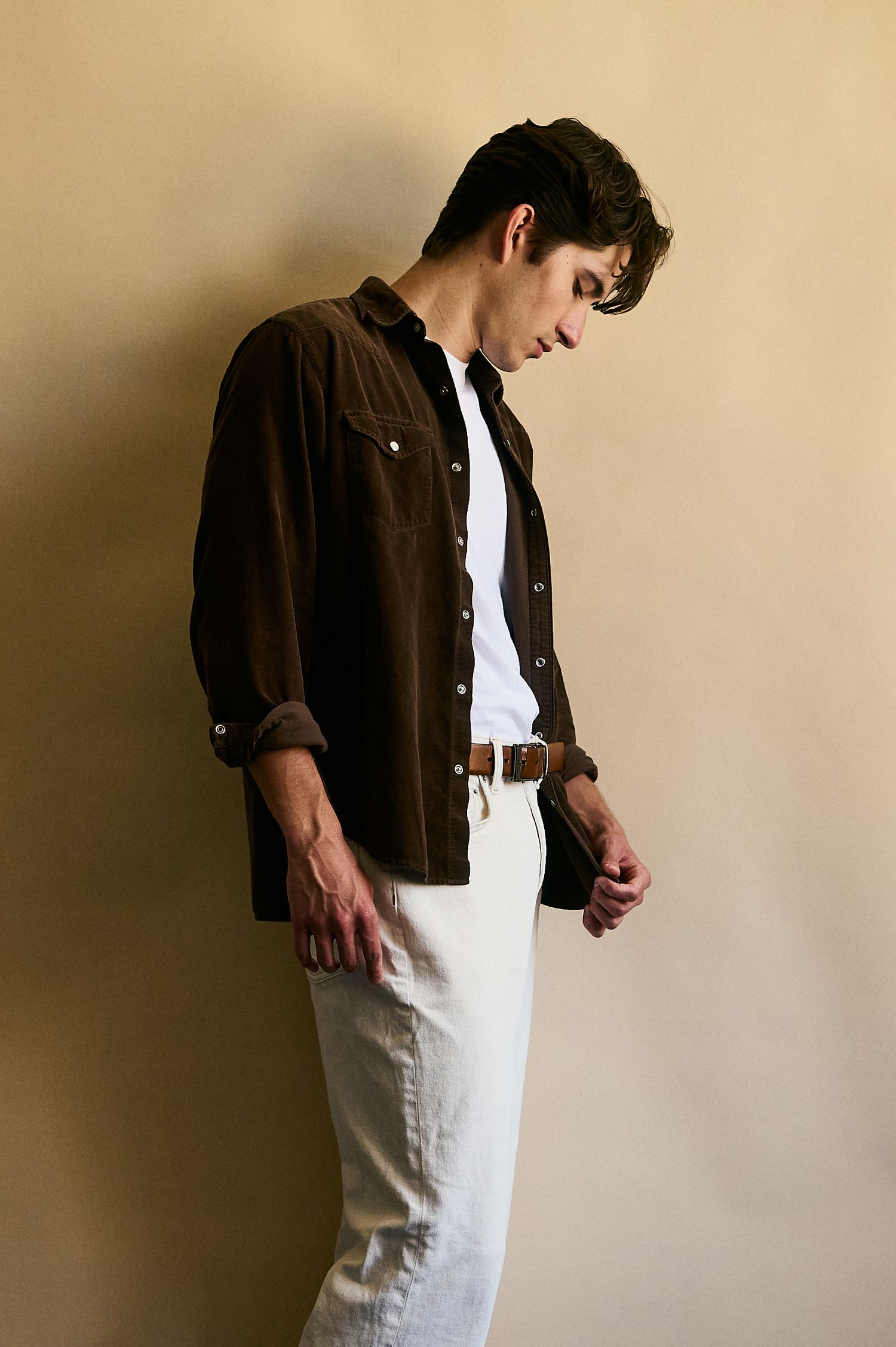 Young man in brown overshirt, white tee, and white trousers with brown belt, posing against beige wall.