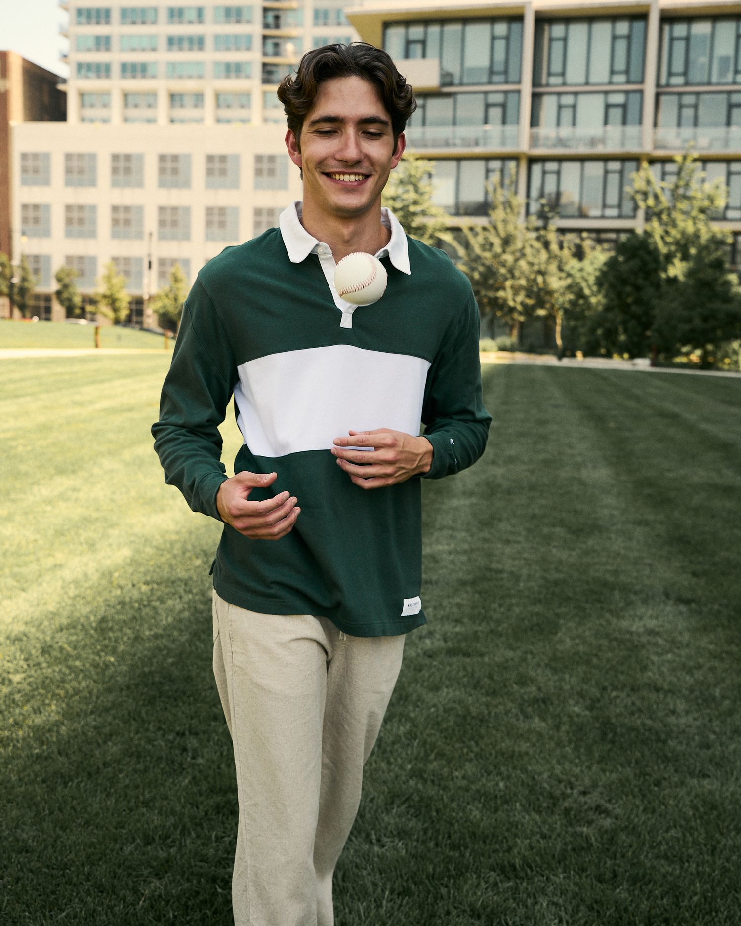 Young man smiling in green and white rugby polo shirt with khaki pants walking on grass outdoors.