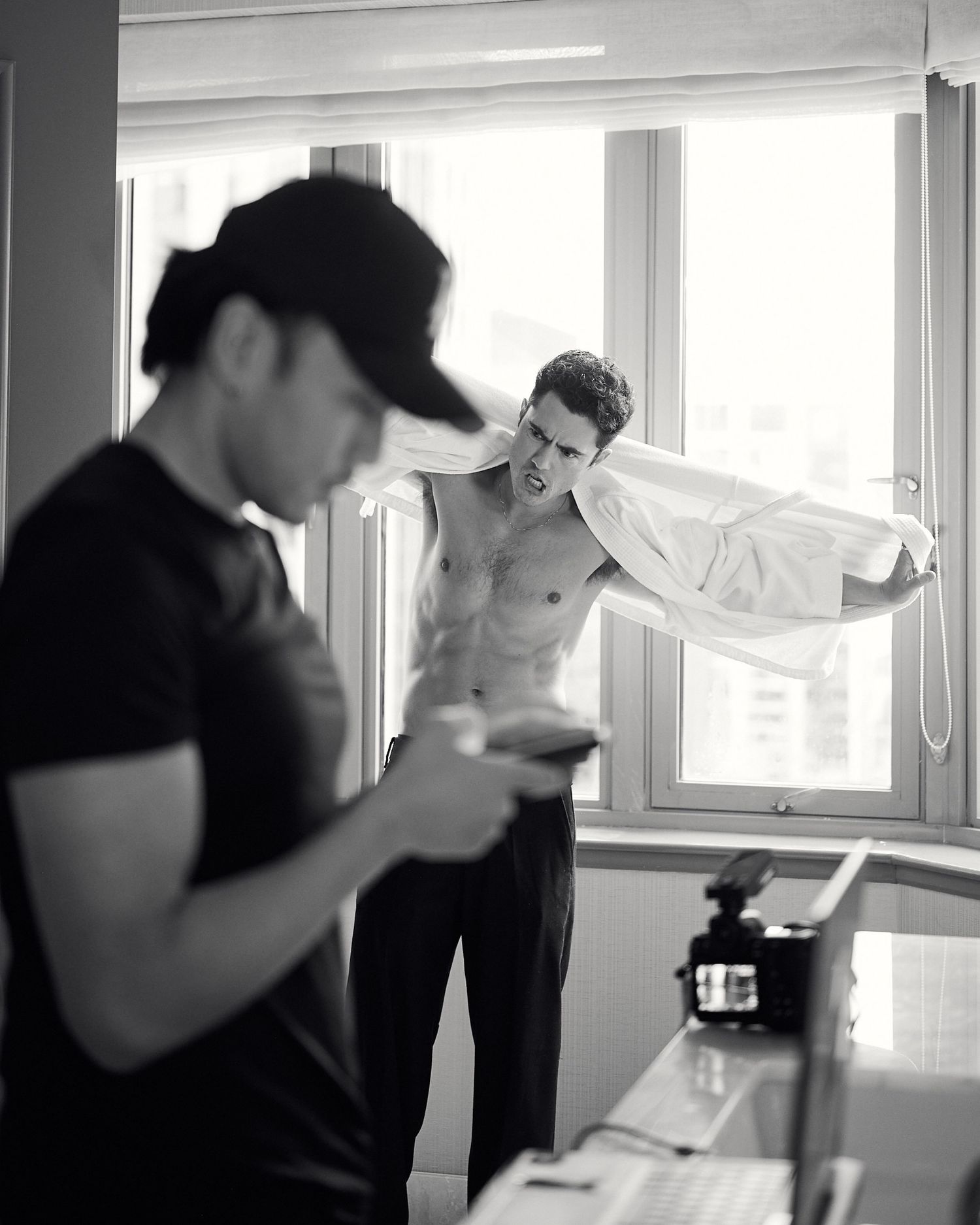 Black and white photo of a shirtless male model putting on a white shirt near a bright window.