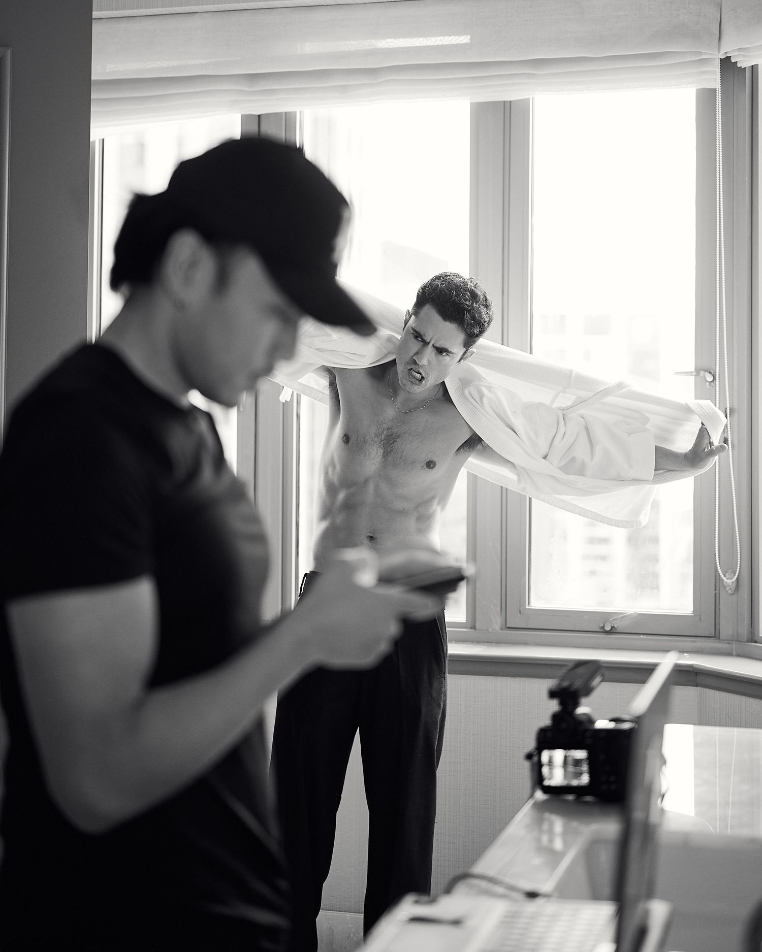 Black and white photo of a shirtless man putting on a white shirt near a window during a photoshoot.