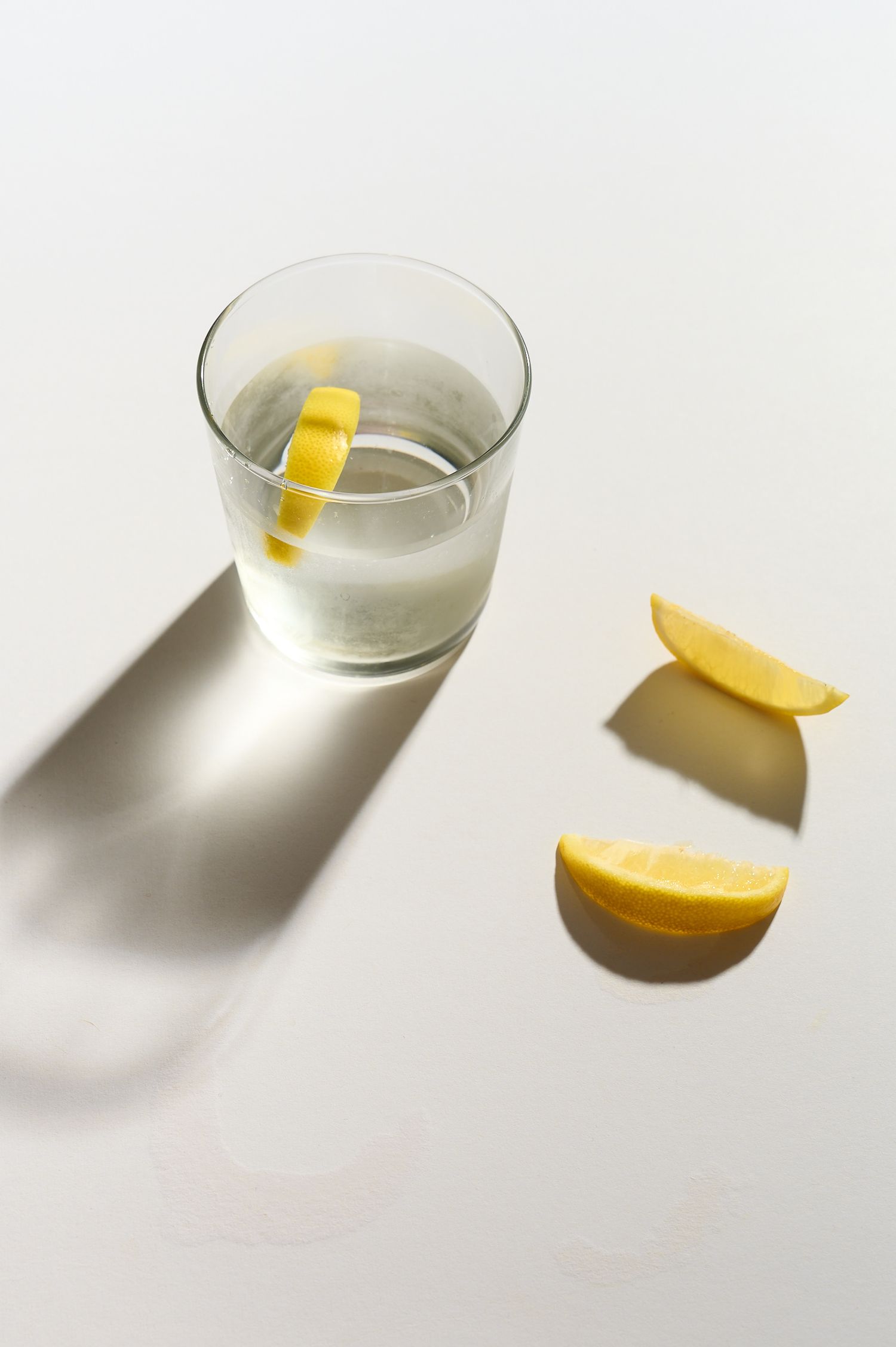 Glass of water with lemon slice on white surface, two lemon wedges beside it casting long shadows in bright light.