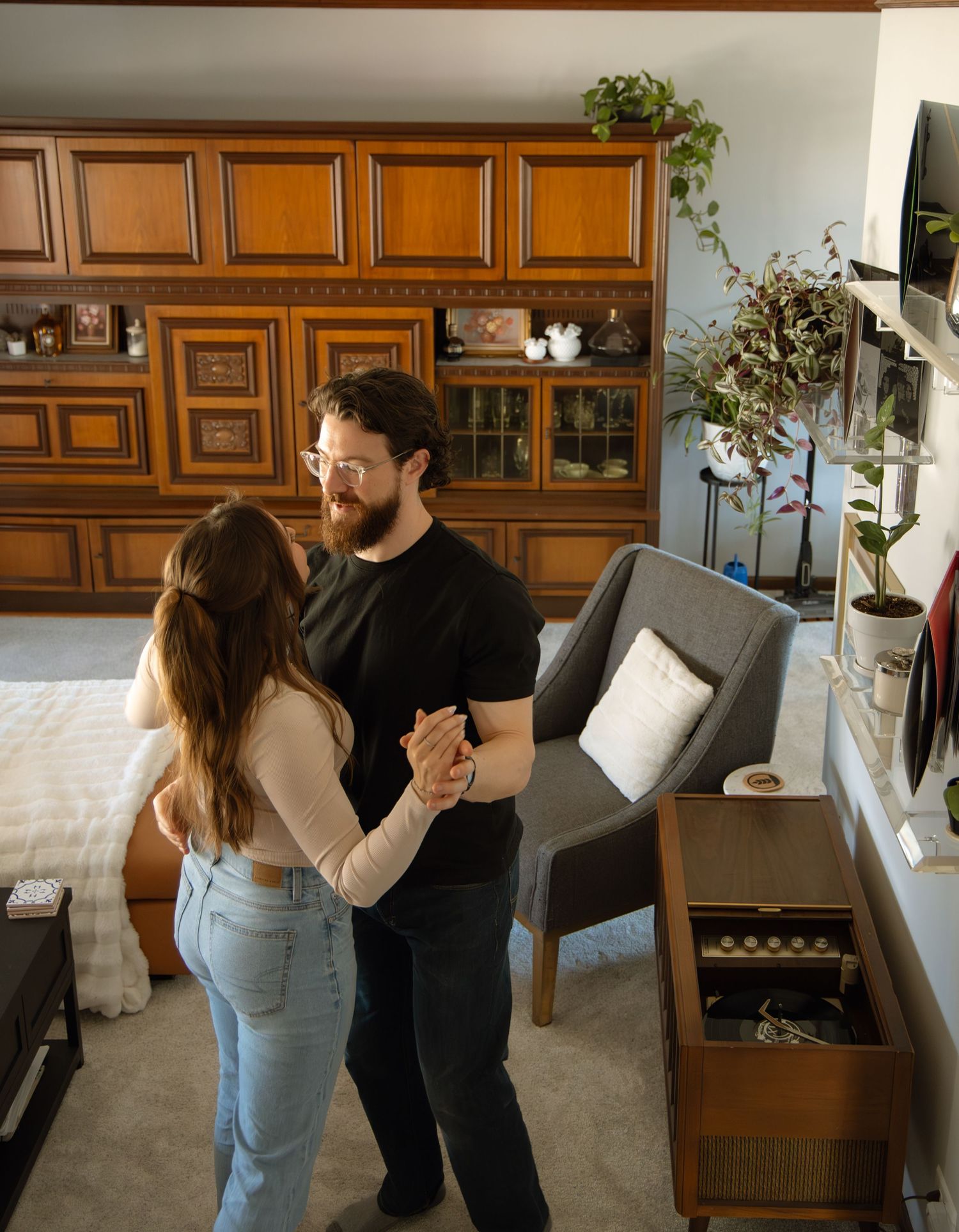 A couple dances together in a cozy living room with wooden cabinetry and houseplants.