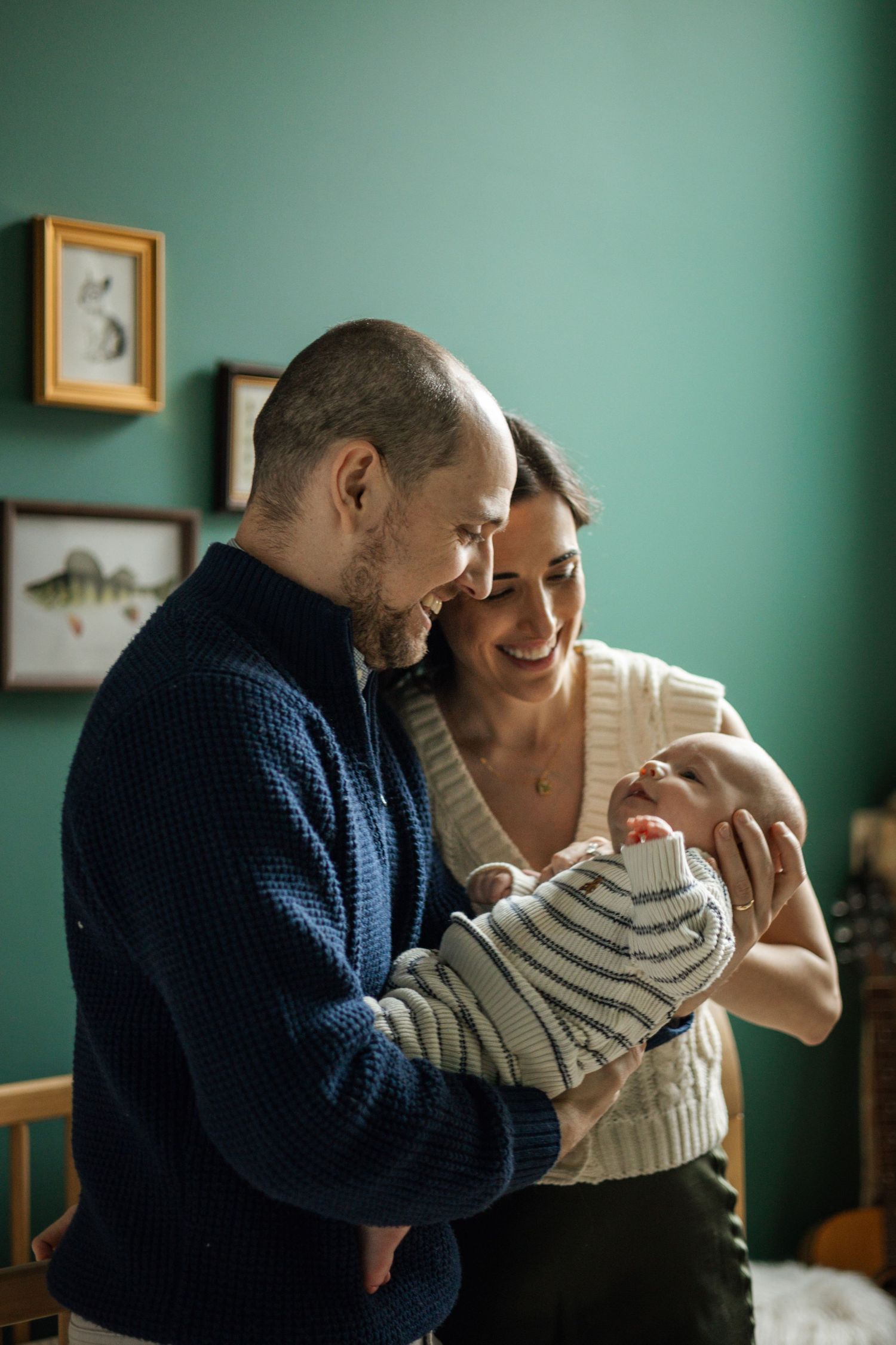 Happy parents smiling and holding their newborn baby in a cozy green nursery room.