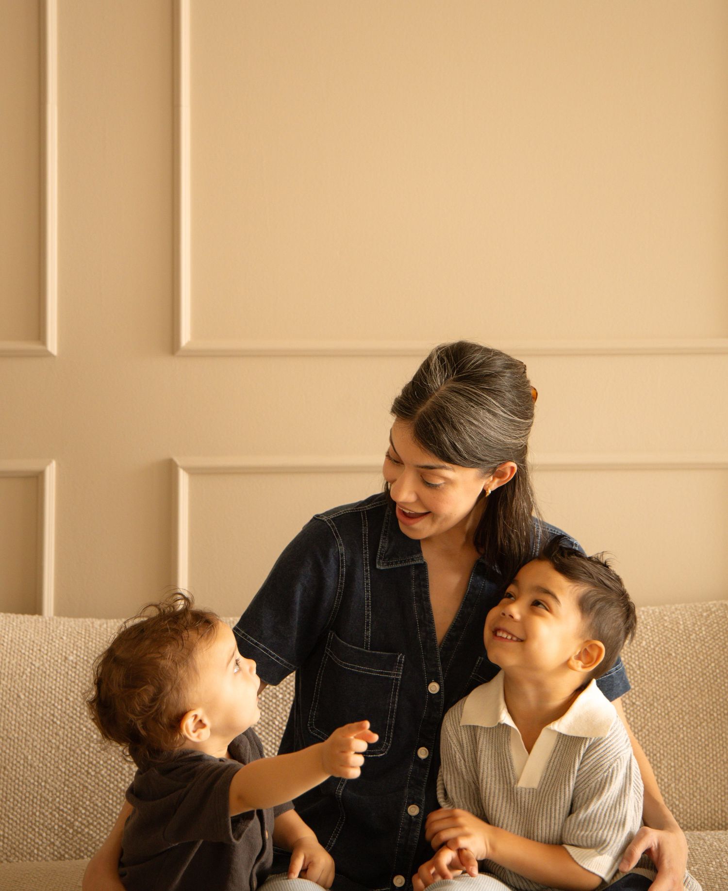 A woman in a denim dress sits on a sofa smiling with two young children in a warm, modern living room.