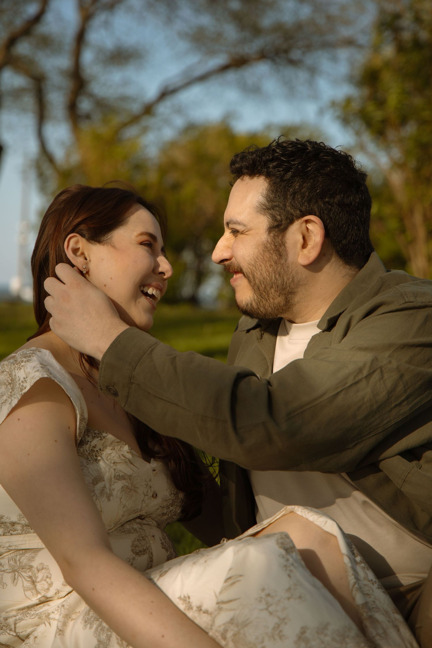 Joyful couple sitting in a park, man tenderly touching womans face as they laugh together.