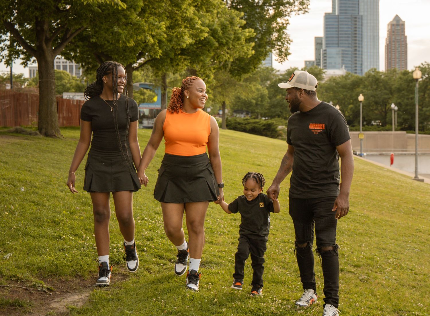 A joyful family of four walks through a green park with city skyline in the background on a warm day.