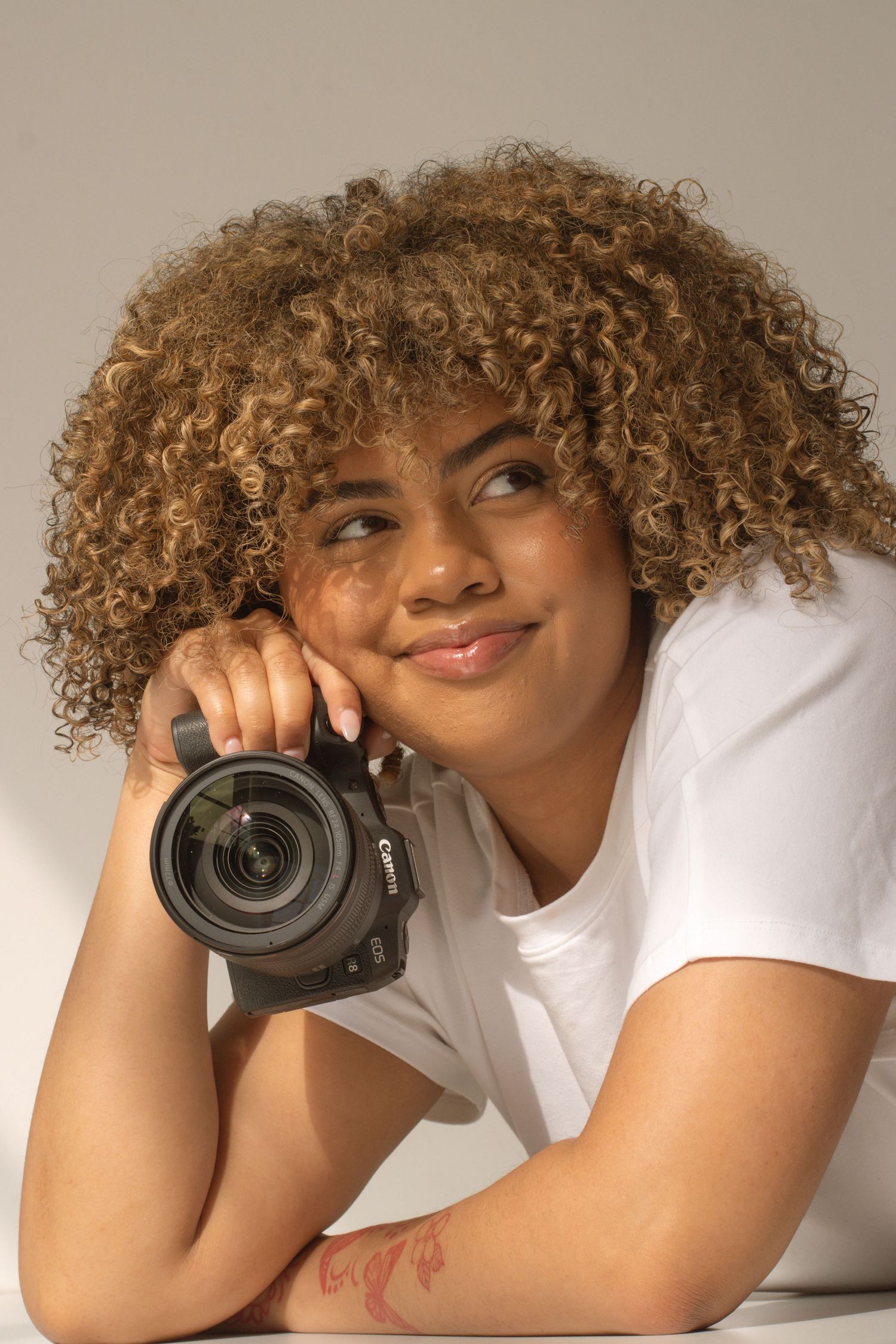 Young woman with curly blonde hair holds a Canon DSLR camera, smiling thoughtfully in a white t-shirt against a neutral background.