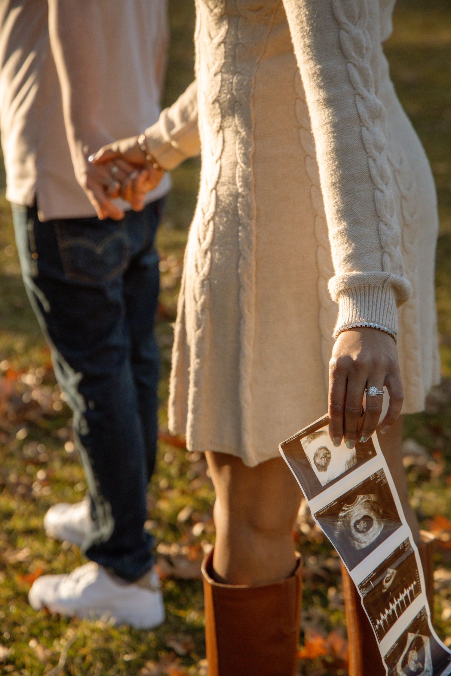 Couple holding hands outdoors in autumn, woman in cream sweater dress holding ultrasound photos.