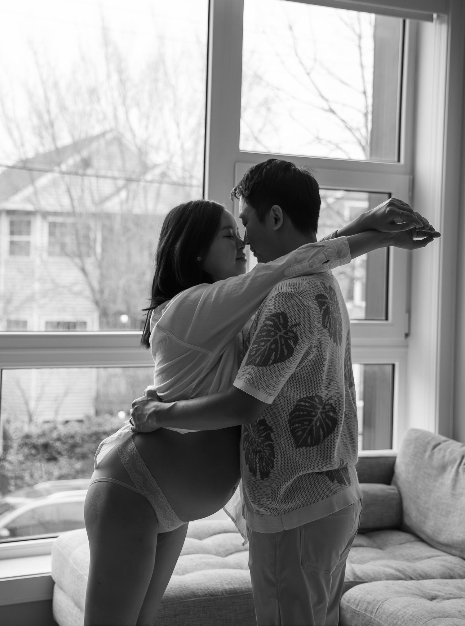 Black and white photo of pregnant woman and partner sharing a kiss by a bright window.