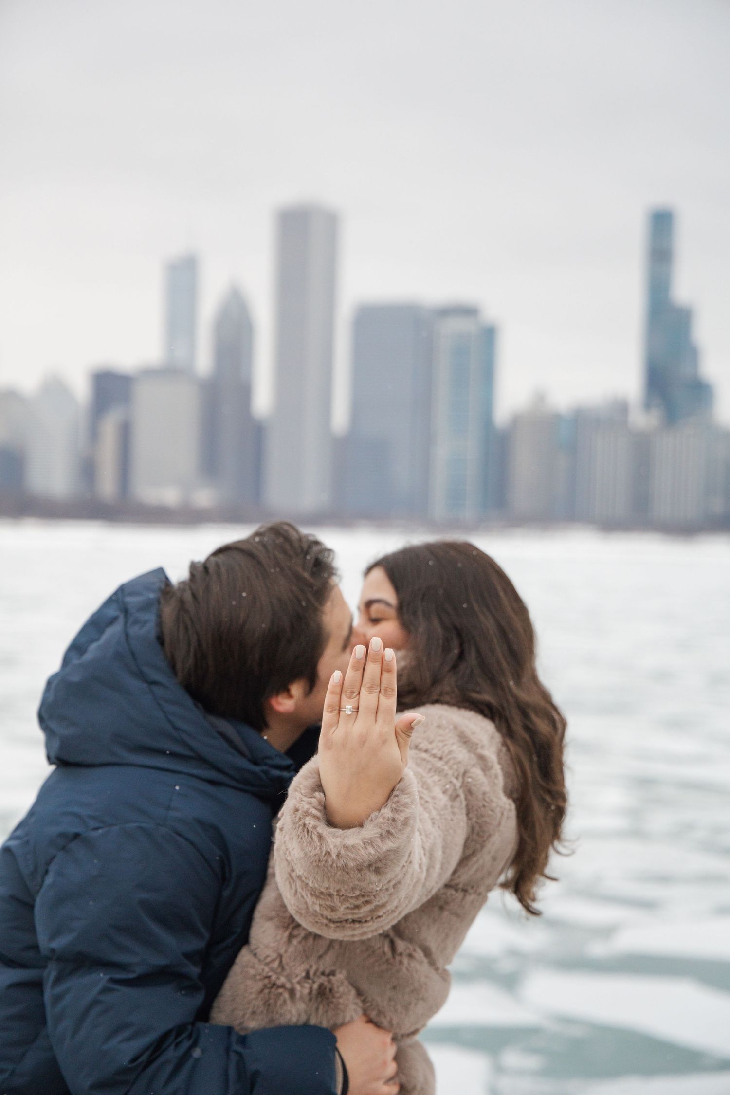 Engaged couple kissing in winter with Chicago skyline backdrop, woman showing diamond ring.