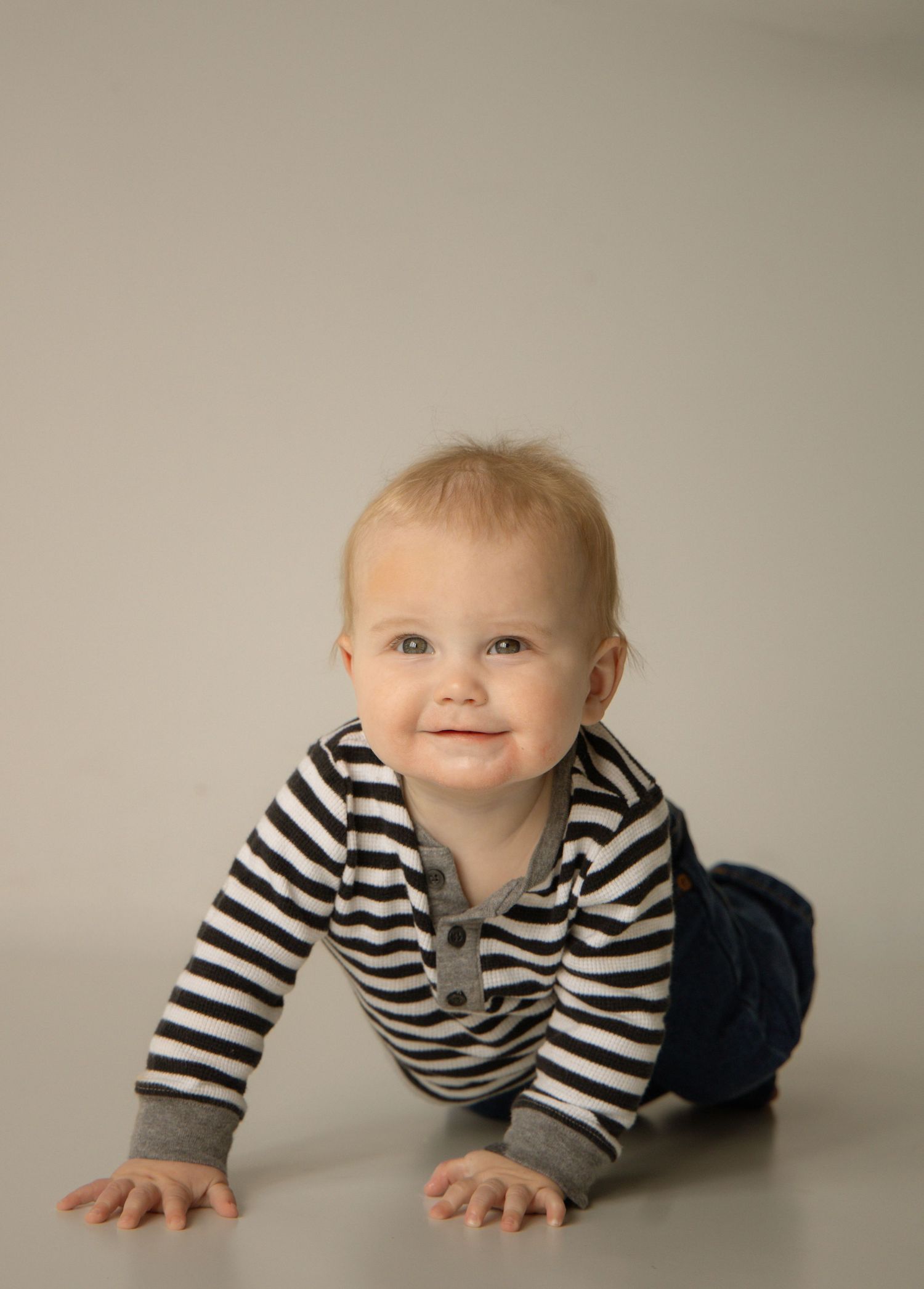 An adorable baby in a black and white striped onesie crawls and smiles on a light gray studio floor.