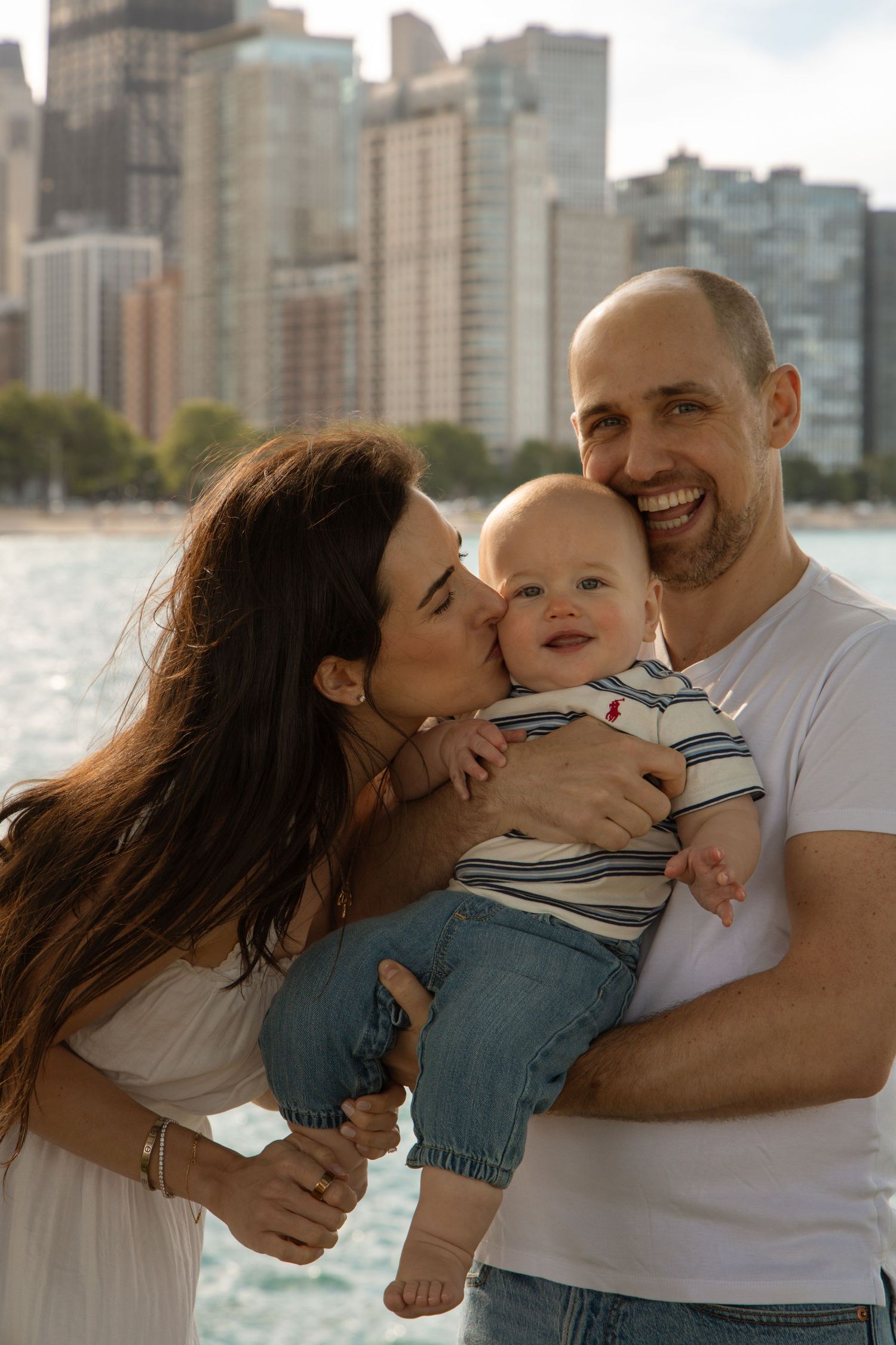 Happy family with mother kissing baby held by smiling father, with city skyline and lake in background.