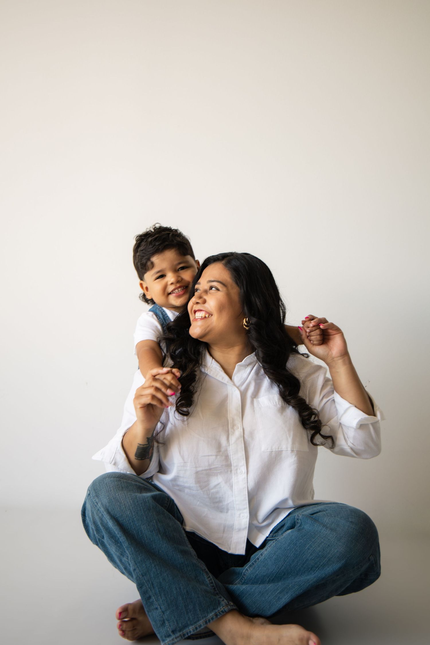 A smiling woman in white shirt and jeans sits cross-legged while a young child hugs her from behind.