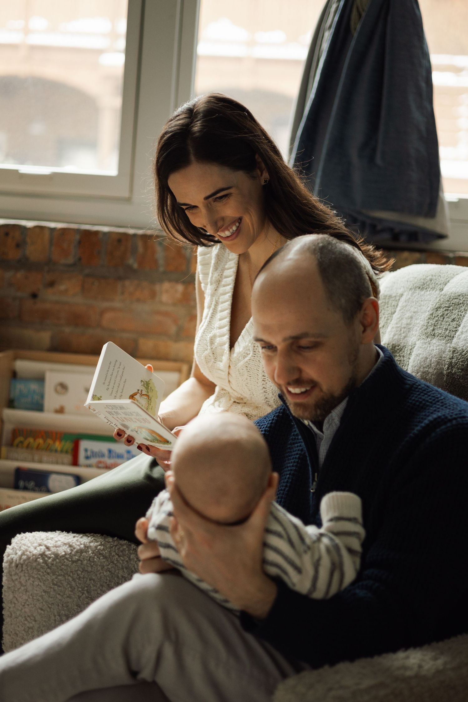 Happy parents smile while reading a book to their newborn baby together in a warmly lit nursery room.