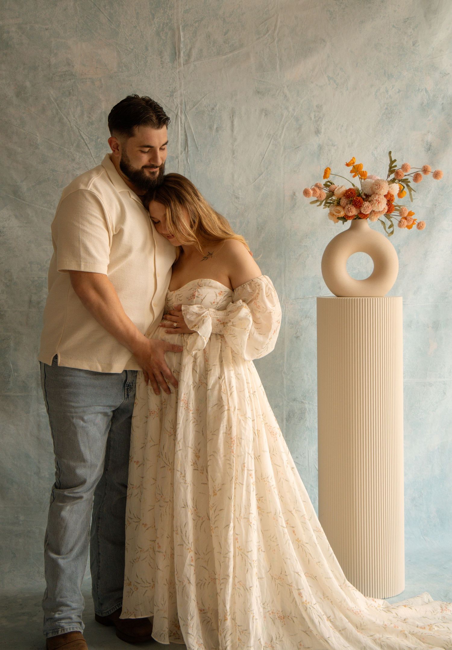 Couple in maternity photo shoot, man embracing pregnant woman in lace gown near floral vase.