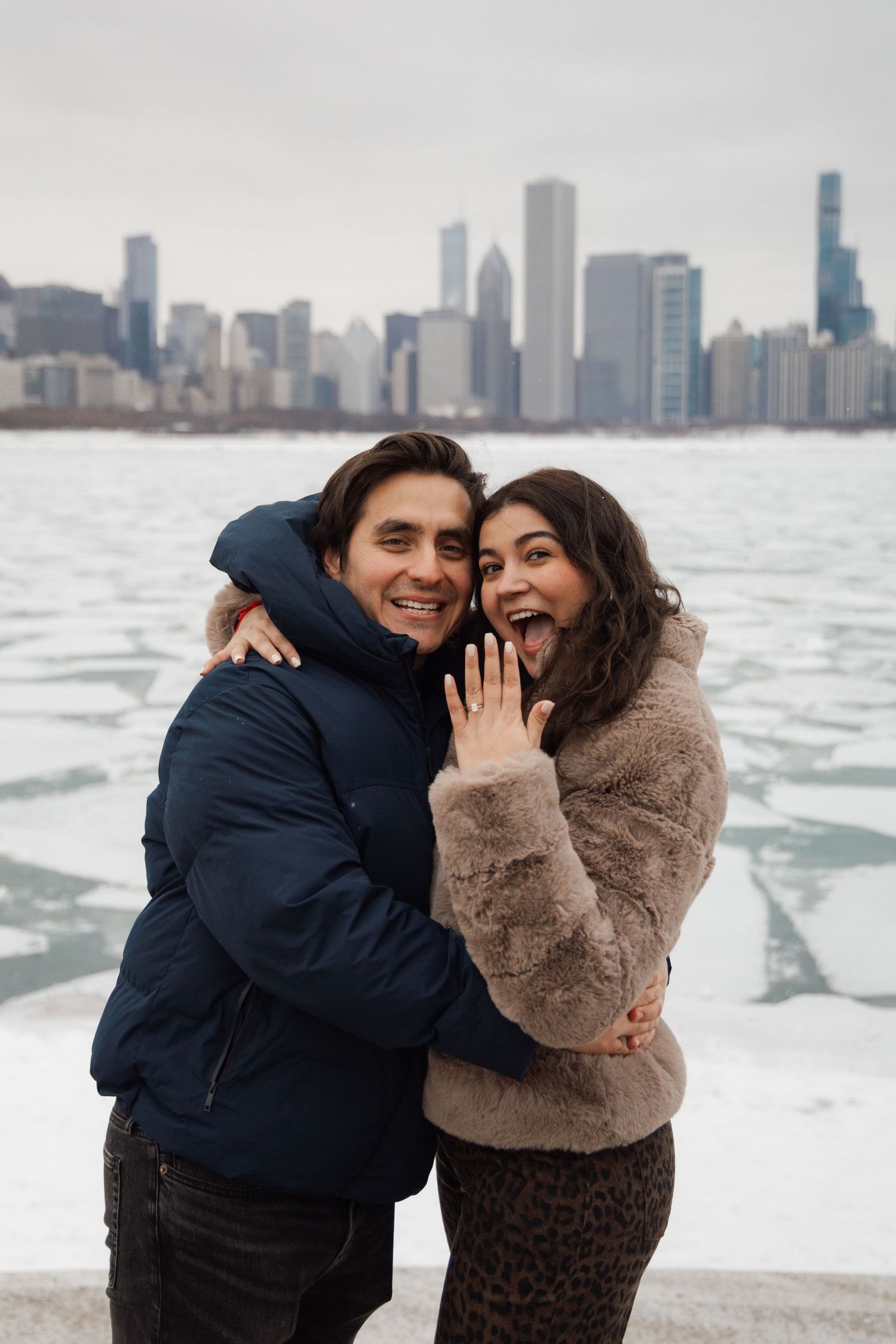 Joyful couple celebrates engagement with Chicago skyline and frozen lake backdrop in winter.