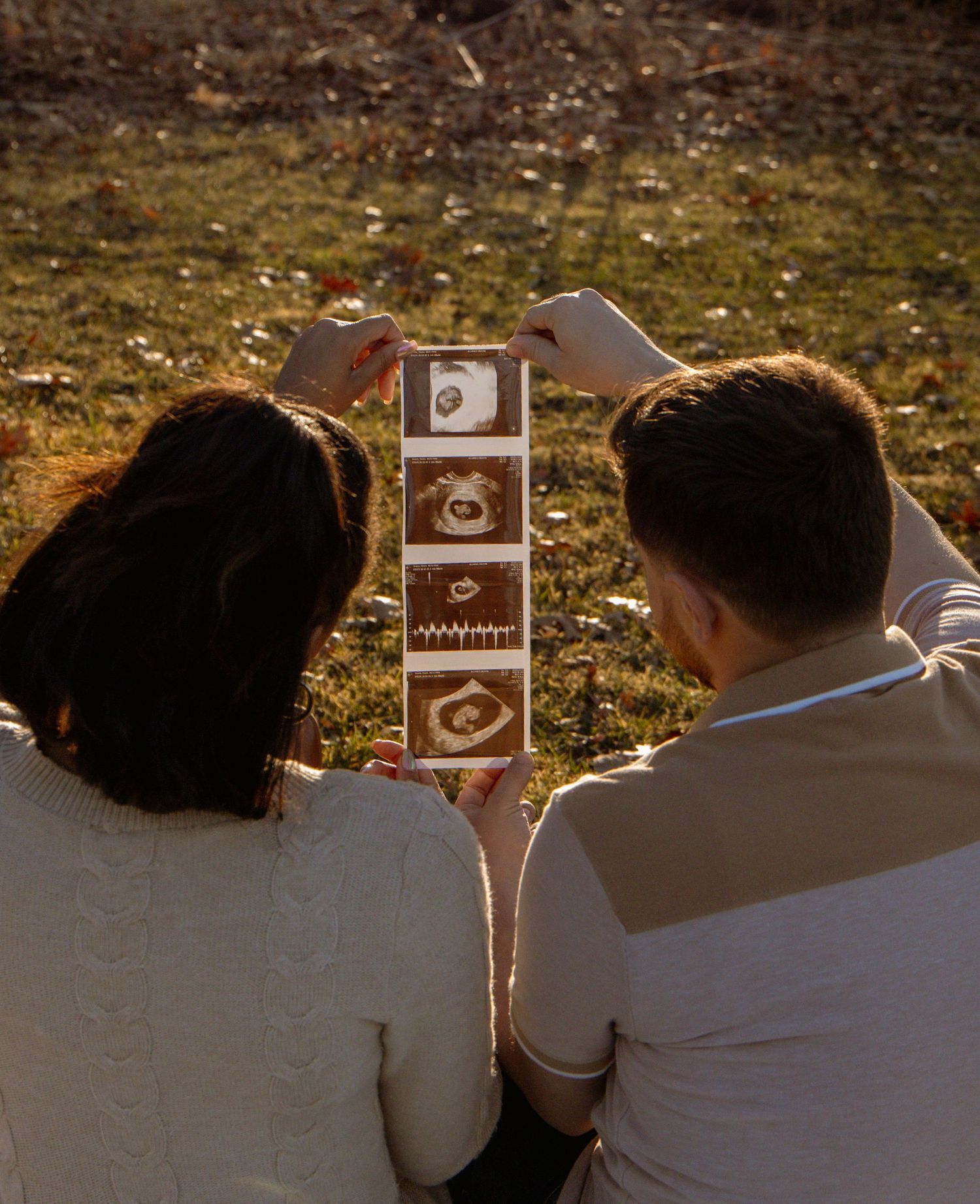 Couple holding ultrasound photos outdoors during golden hour on grass background.