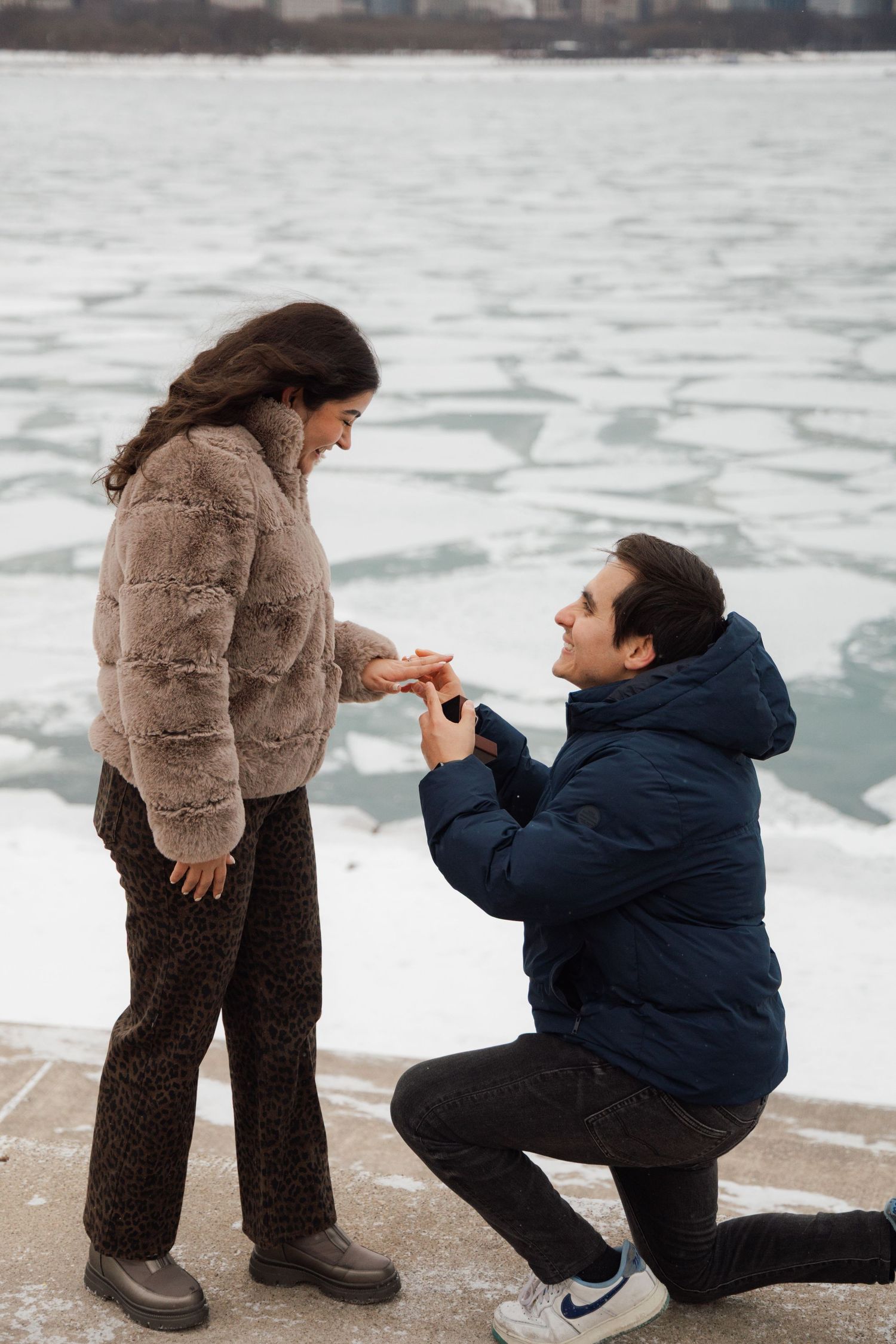 Man kneeling on one knee proposing to woman by frozen river in winter, romantic marriage proposal outdoors.