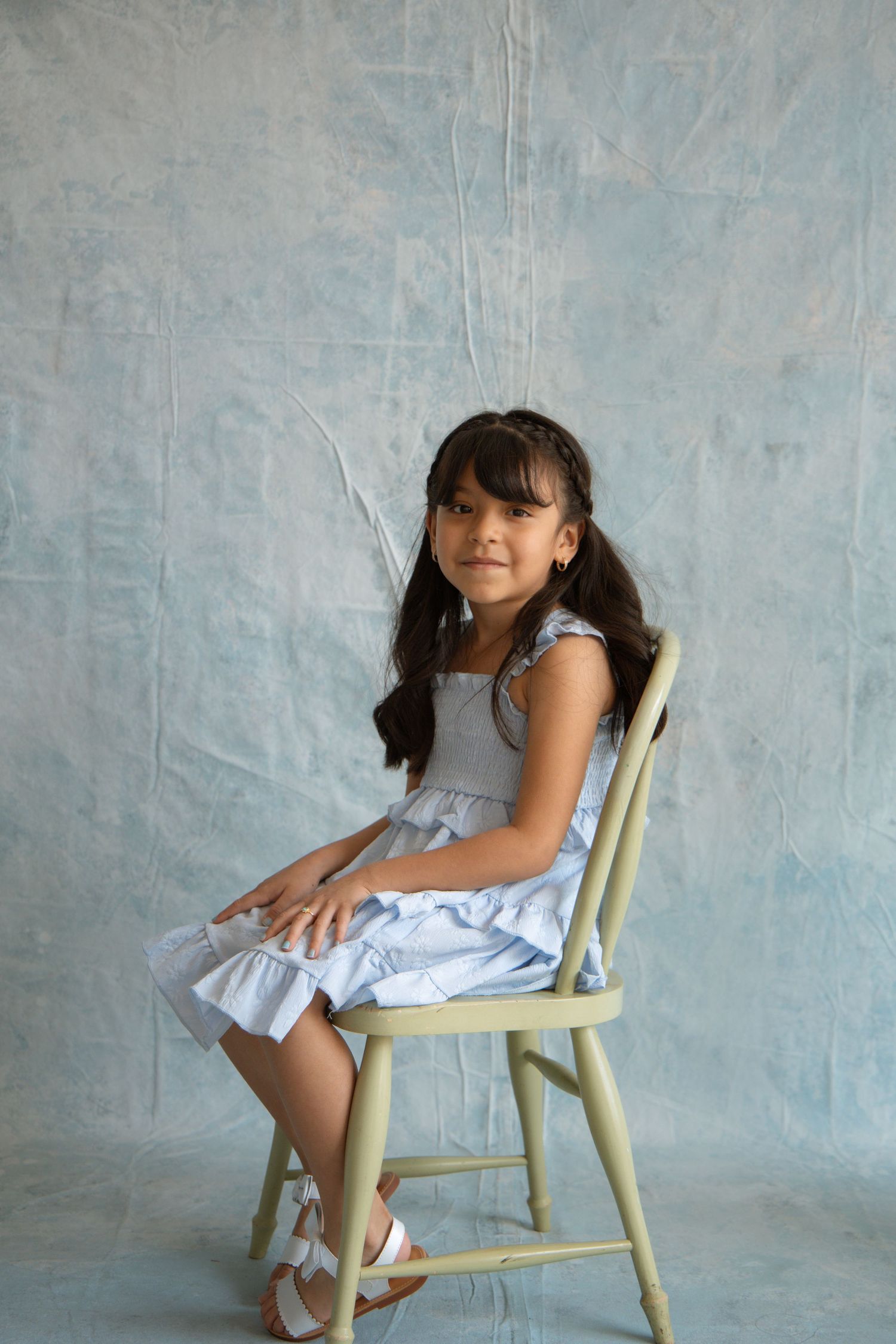 Young girl in a light blue dress sitting on a vintage wooden chair against a textured blue-gray backdrop.