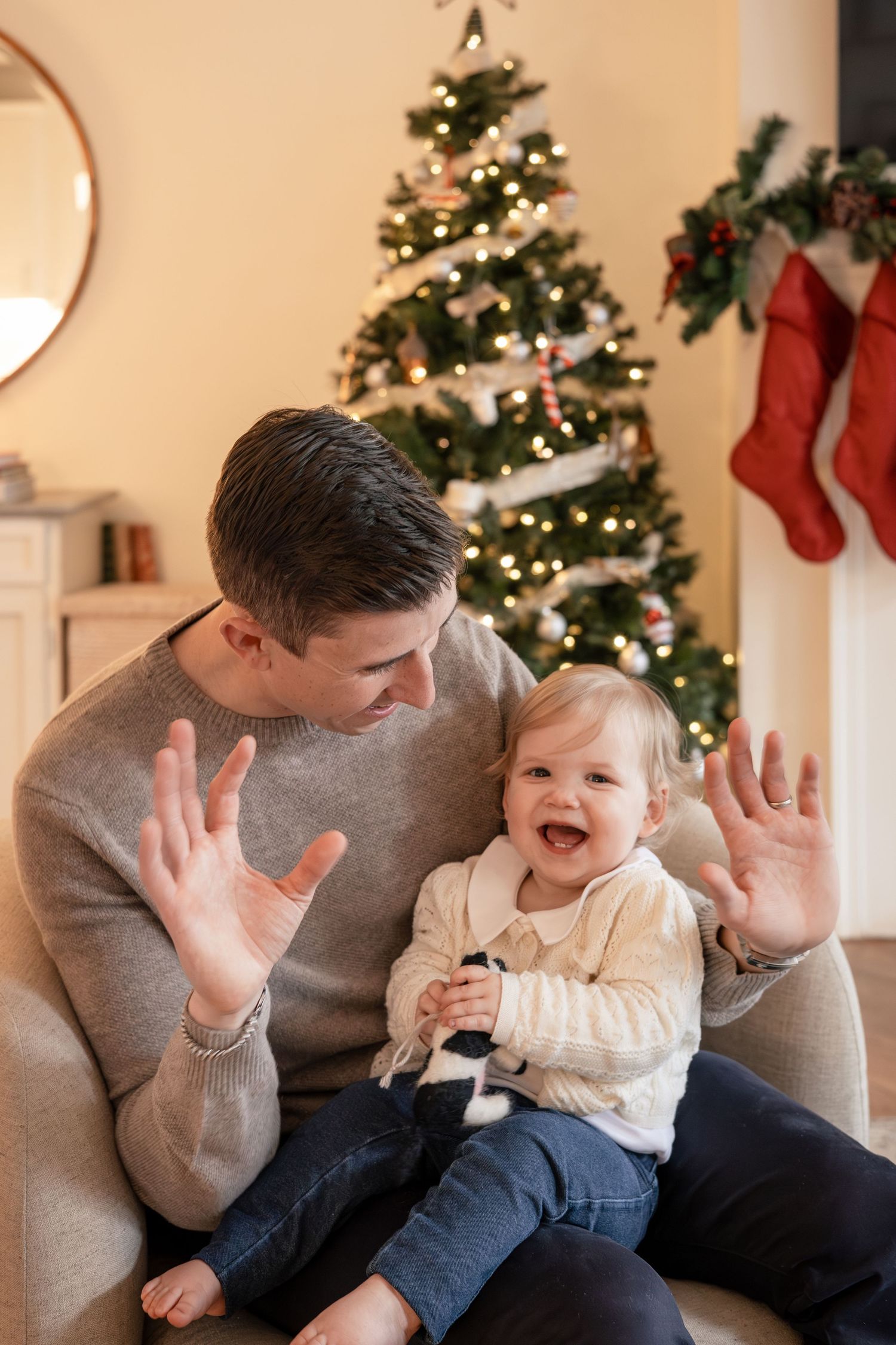 Father and baby laughing together in front of a decorated Christmas tree with stockings hung on fireplace.