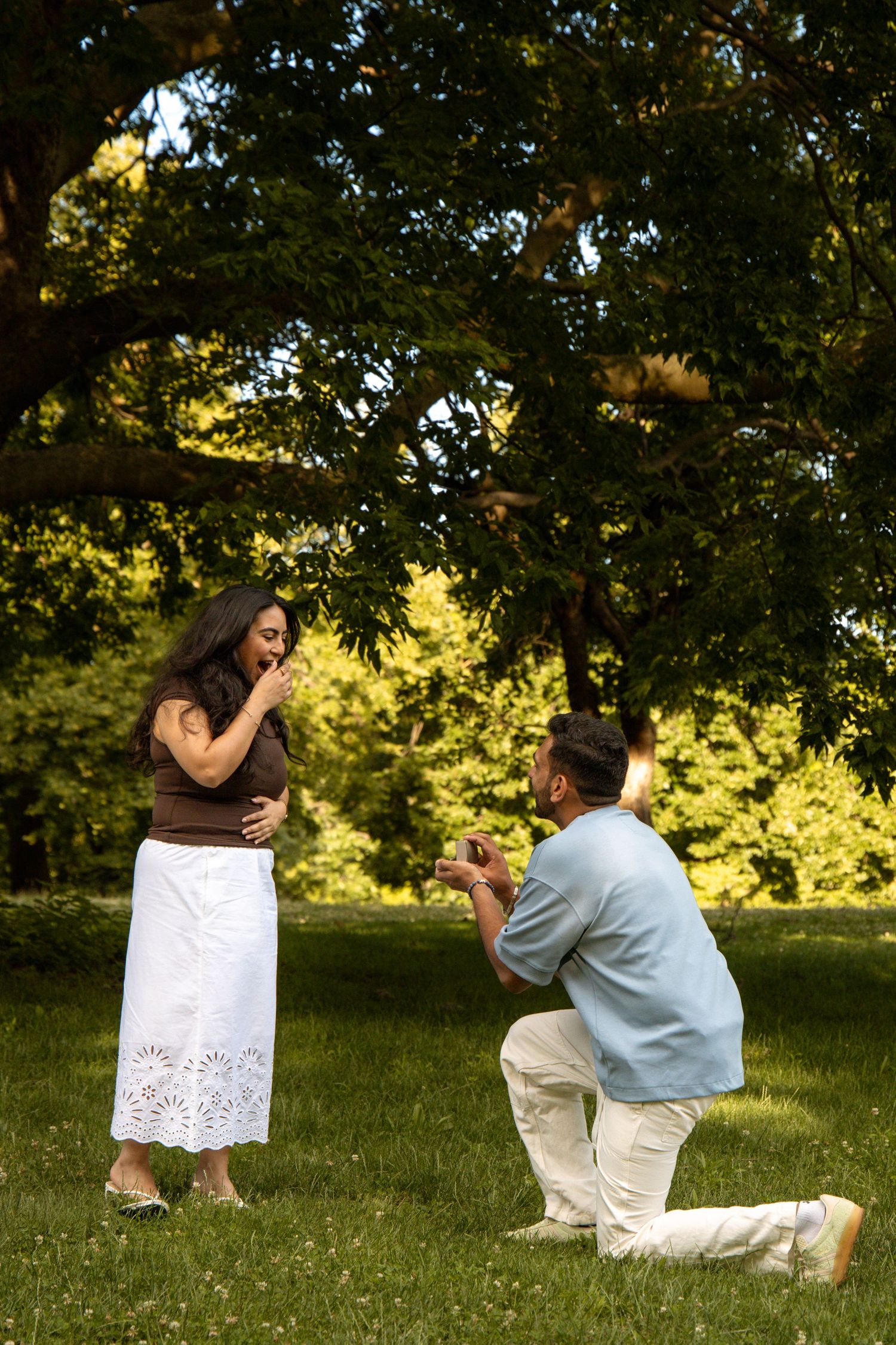 Man kneeling on one knee proposing to woman in a park, holding ring box as she covers her mouth in surprise.