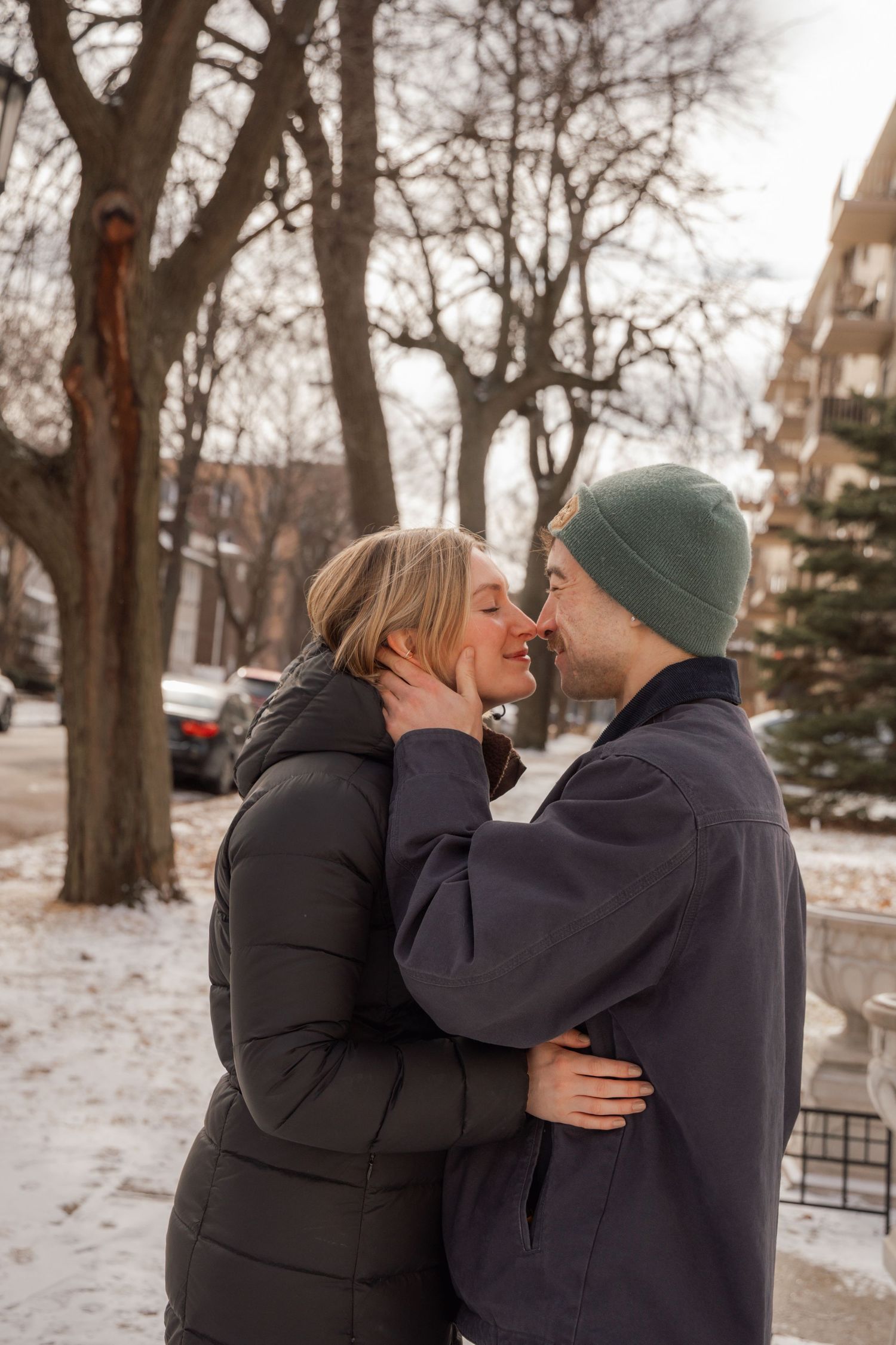 Couple sharing a kiss outdoors in winter, surrounded by snow-covered trees and apartment buildings.