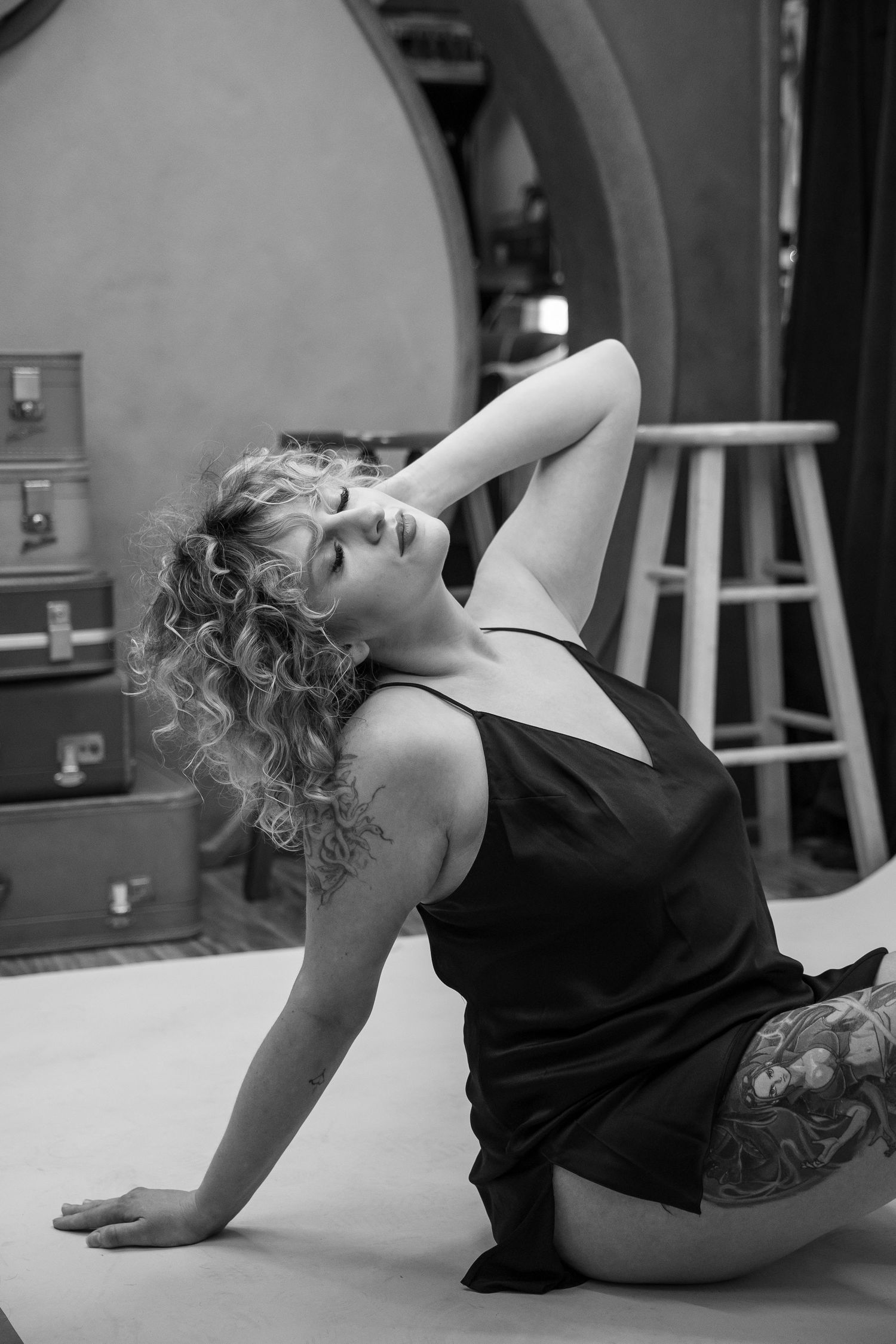 Black and white photo of tattooed blonde woman posing on floor in vintage studio setting.