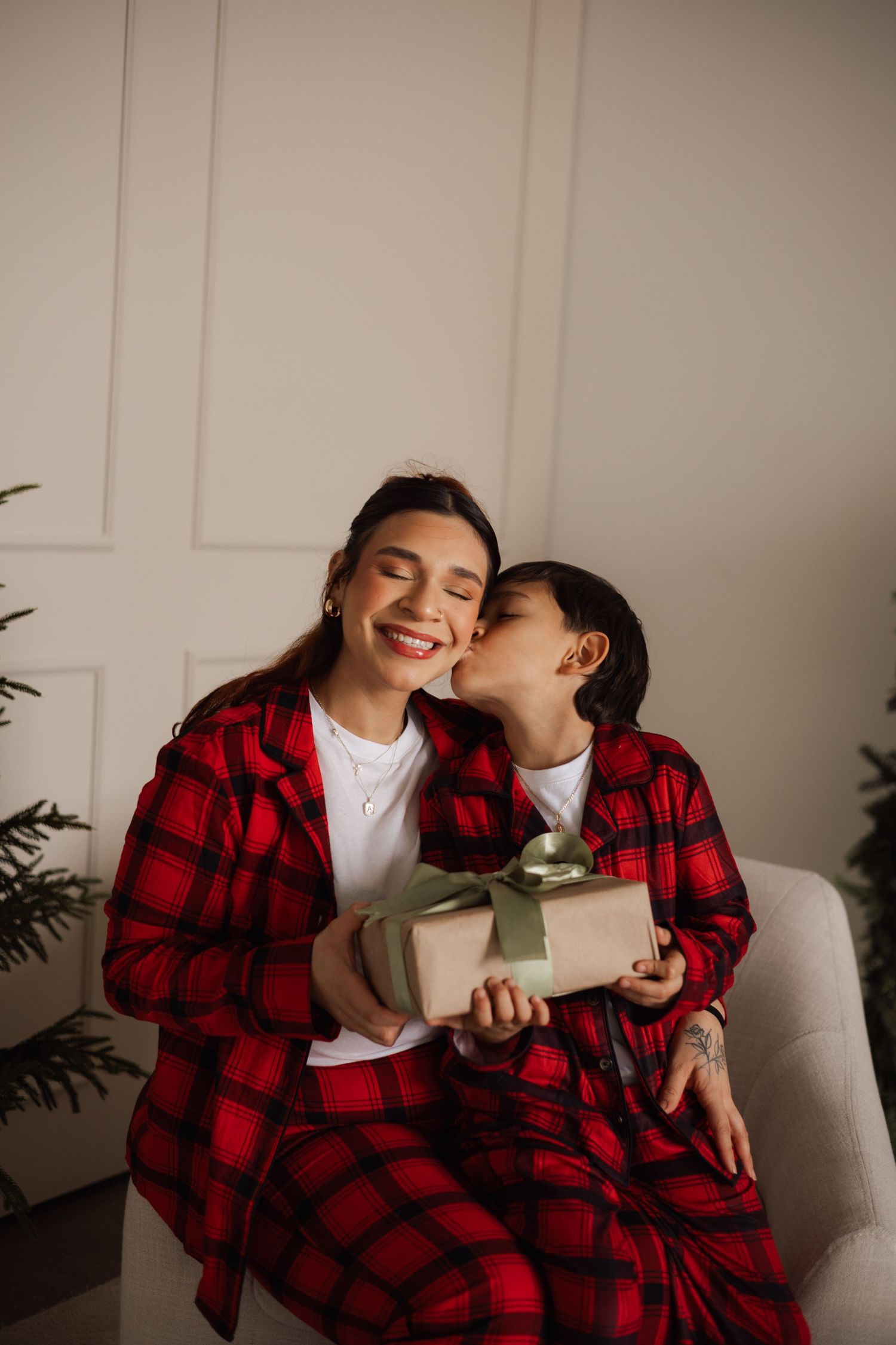 A smiling woman in red plaid pajamas receives a kiss on the cheek while holding a wrapped gift.