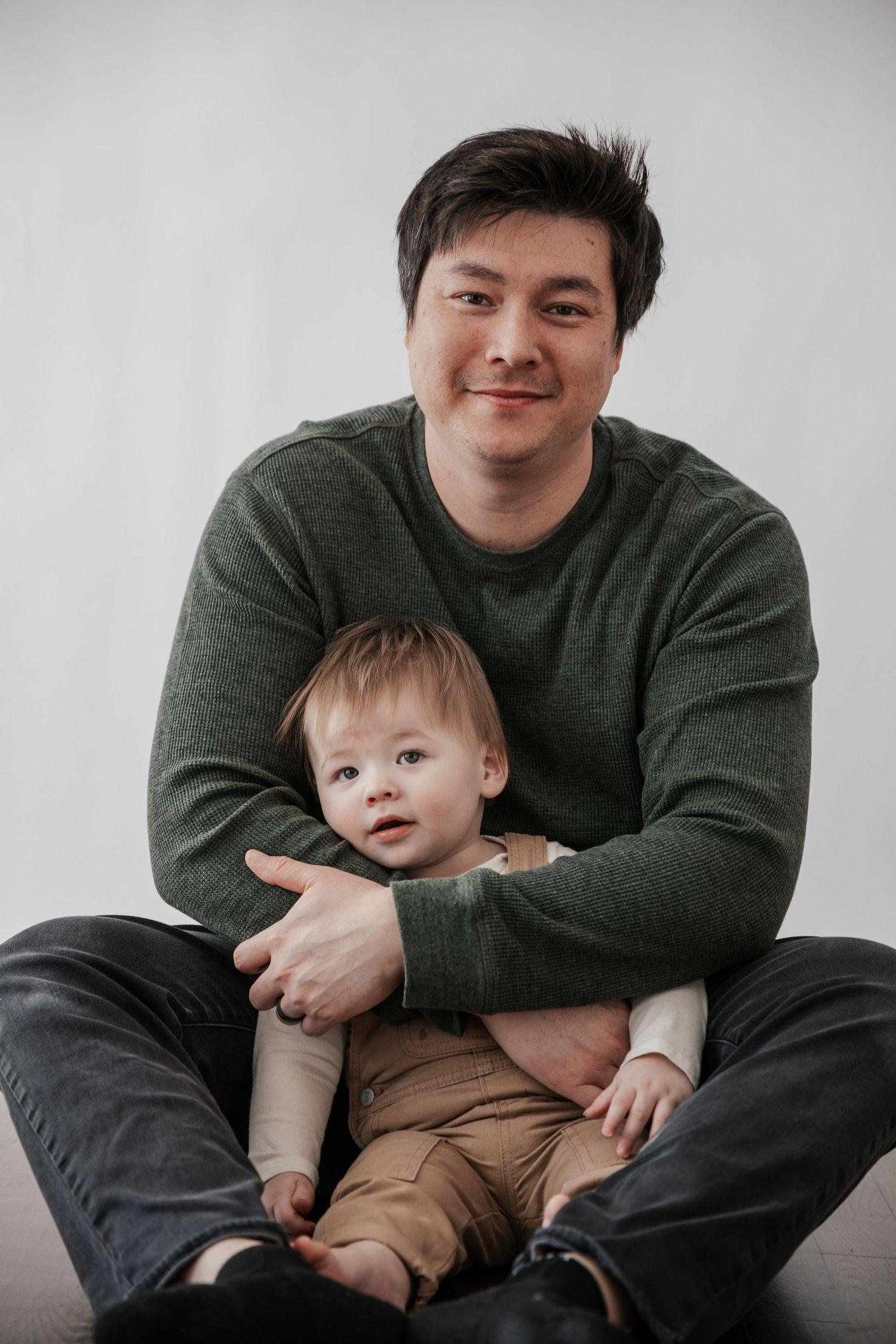 A father in a green sweater sits on the floor cradling his baby in a warm studio portrait.