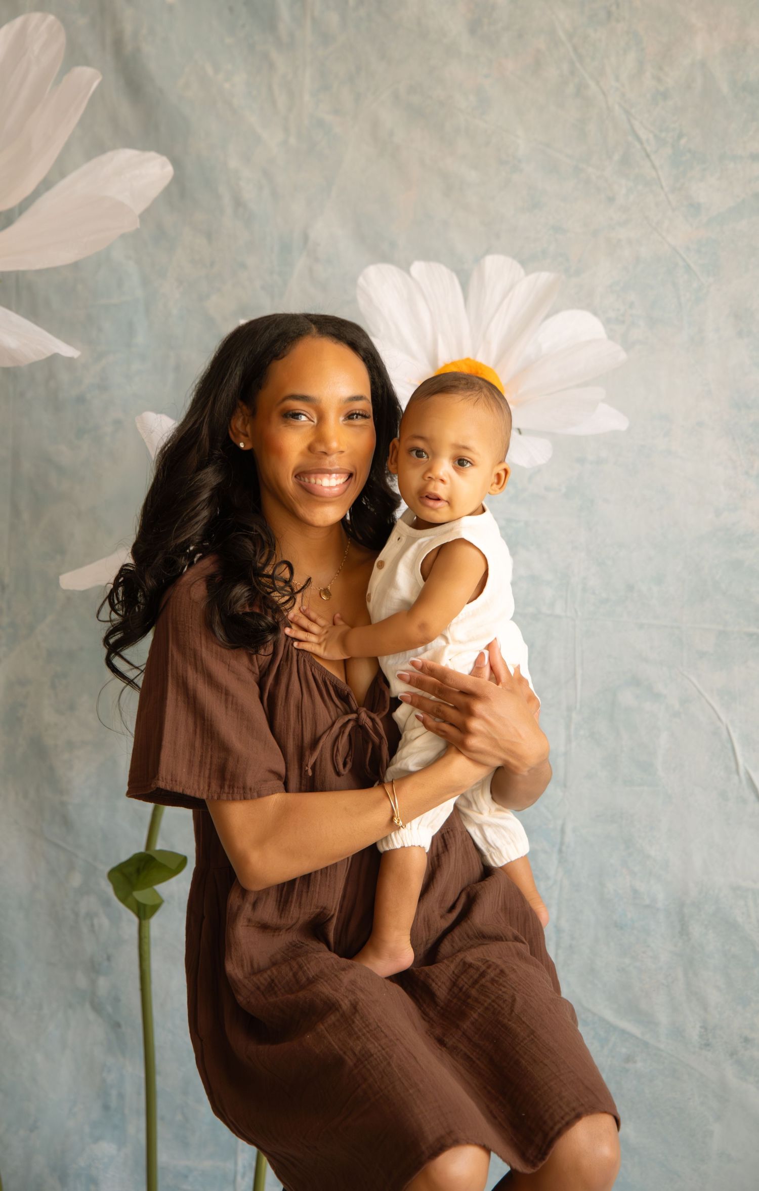 A mother in a brown dress holds her baby with a yellow bow in front of a floral backdrop.