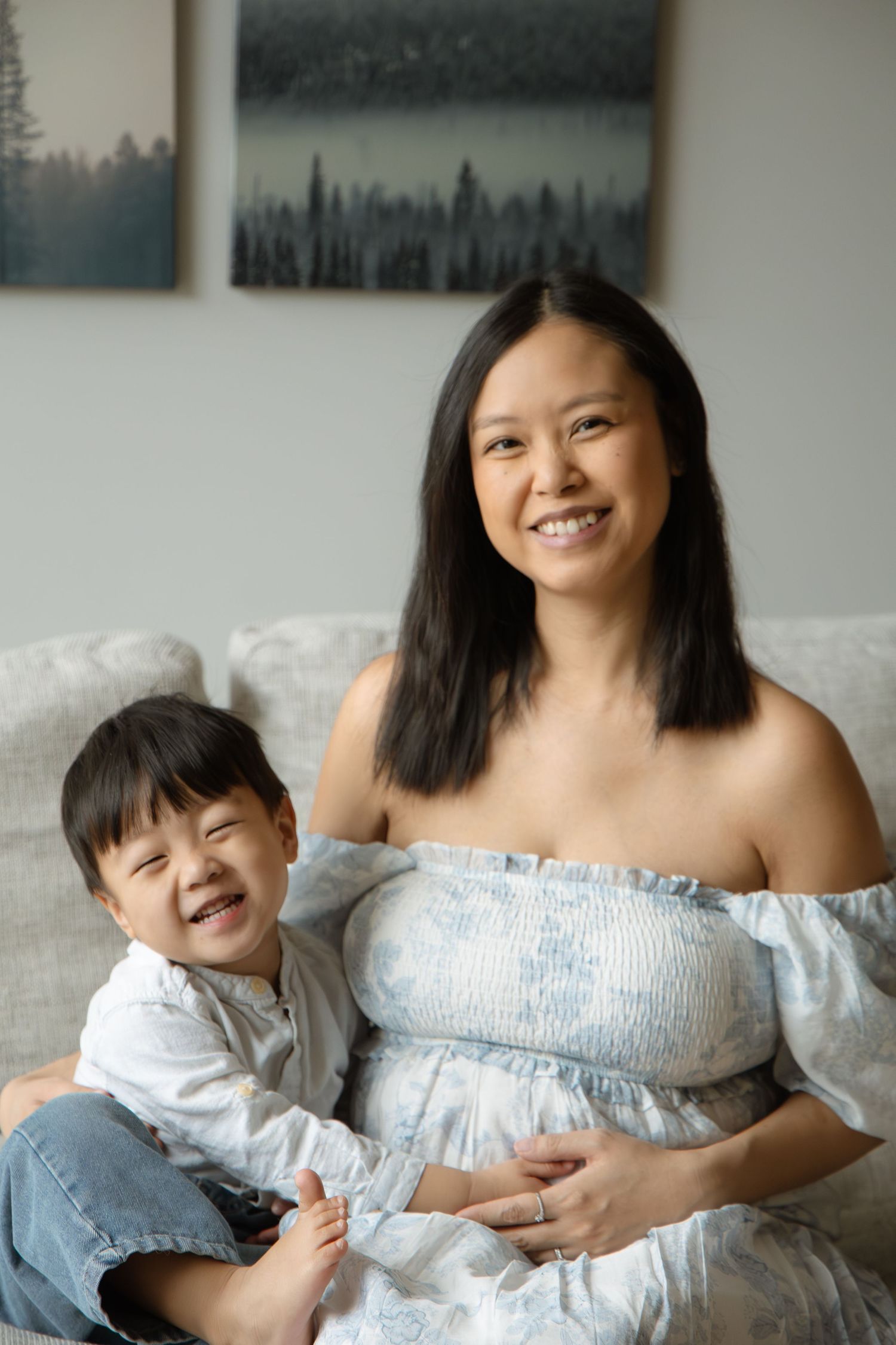 Smiling pregnant Asian woman in blue off-shoulder dress sits on sofa with laughing young son.