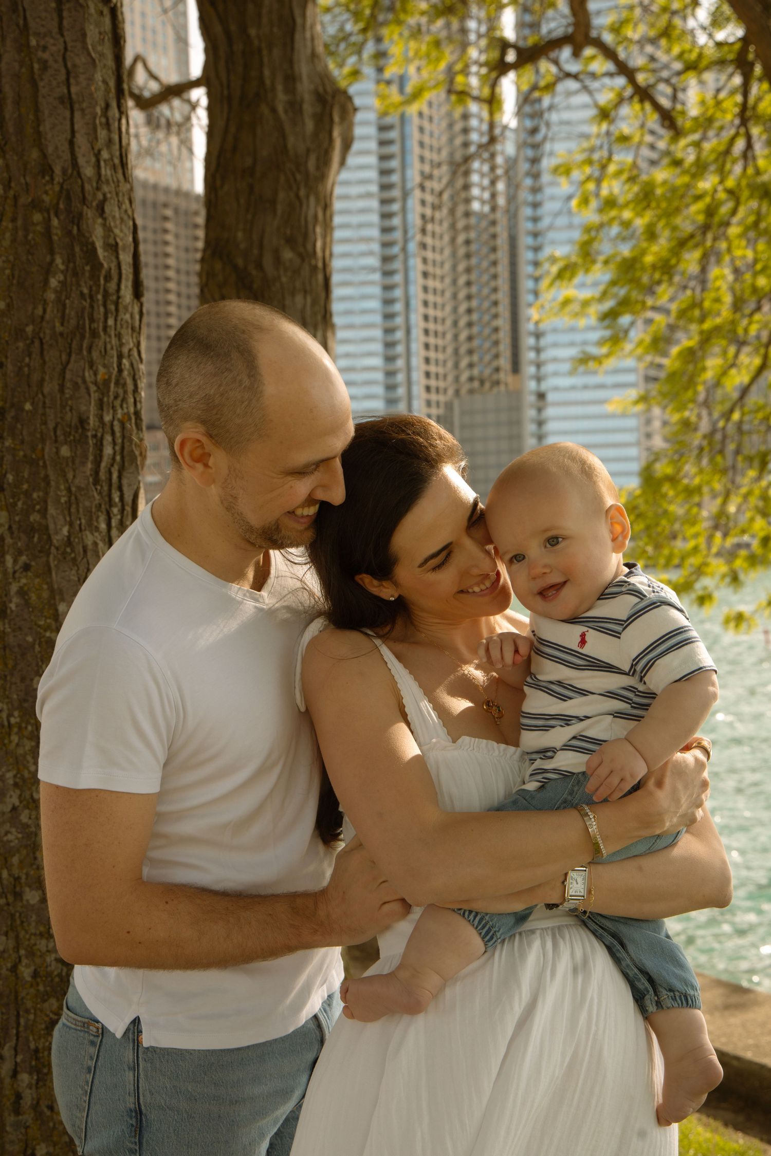 A happy young family with a bald father, dark-haired mother in white dress, and smiling baby boy outdoors by a waterfront.