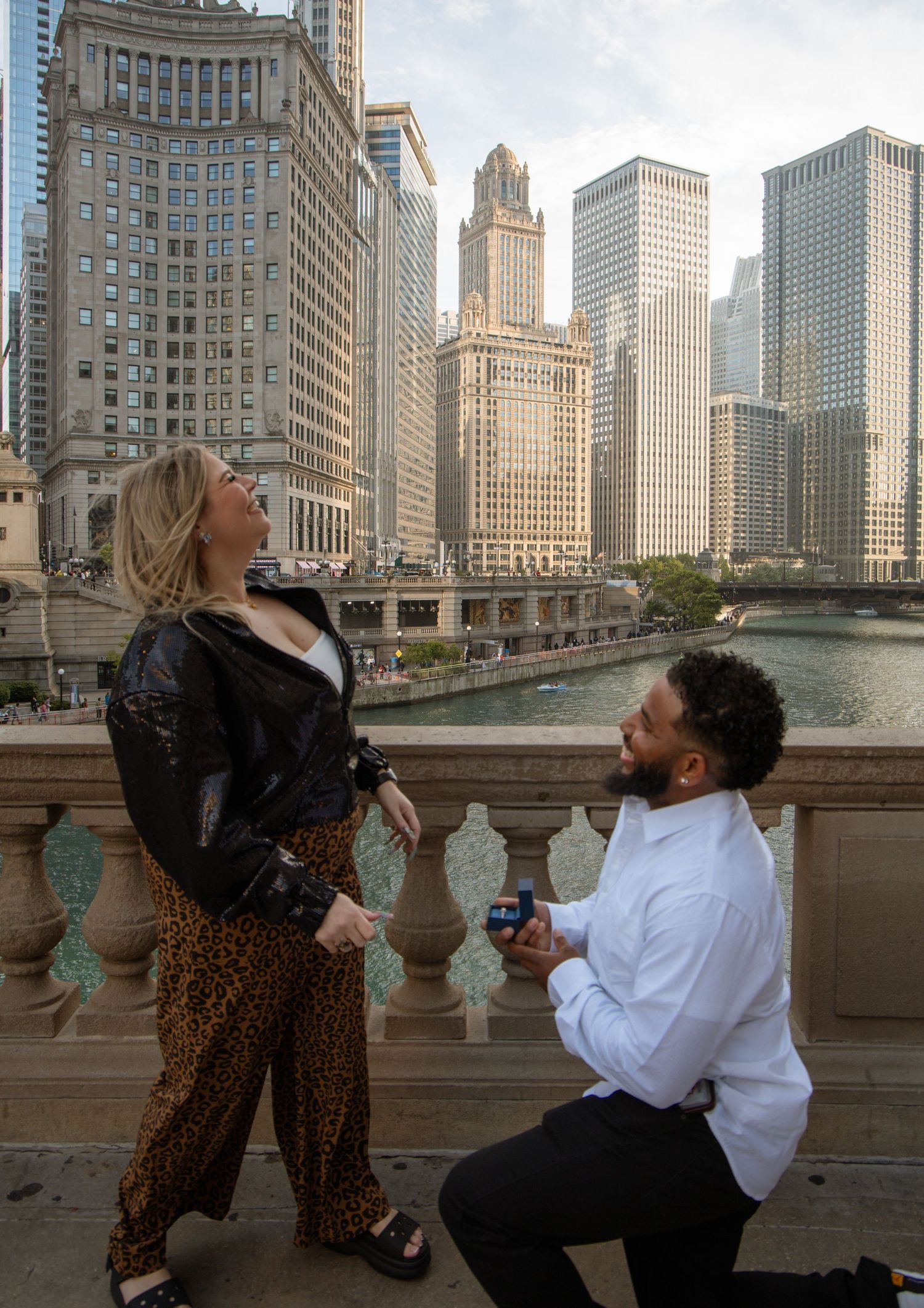 Man proposes on one knee to woman on Chicago Riverwalk with stunning city skyline backdrop.