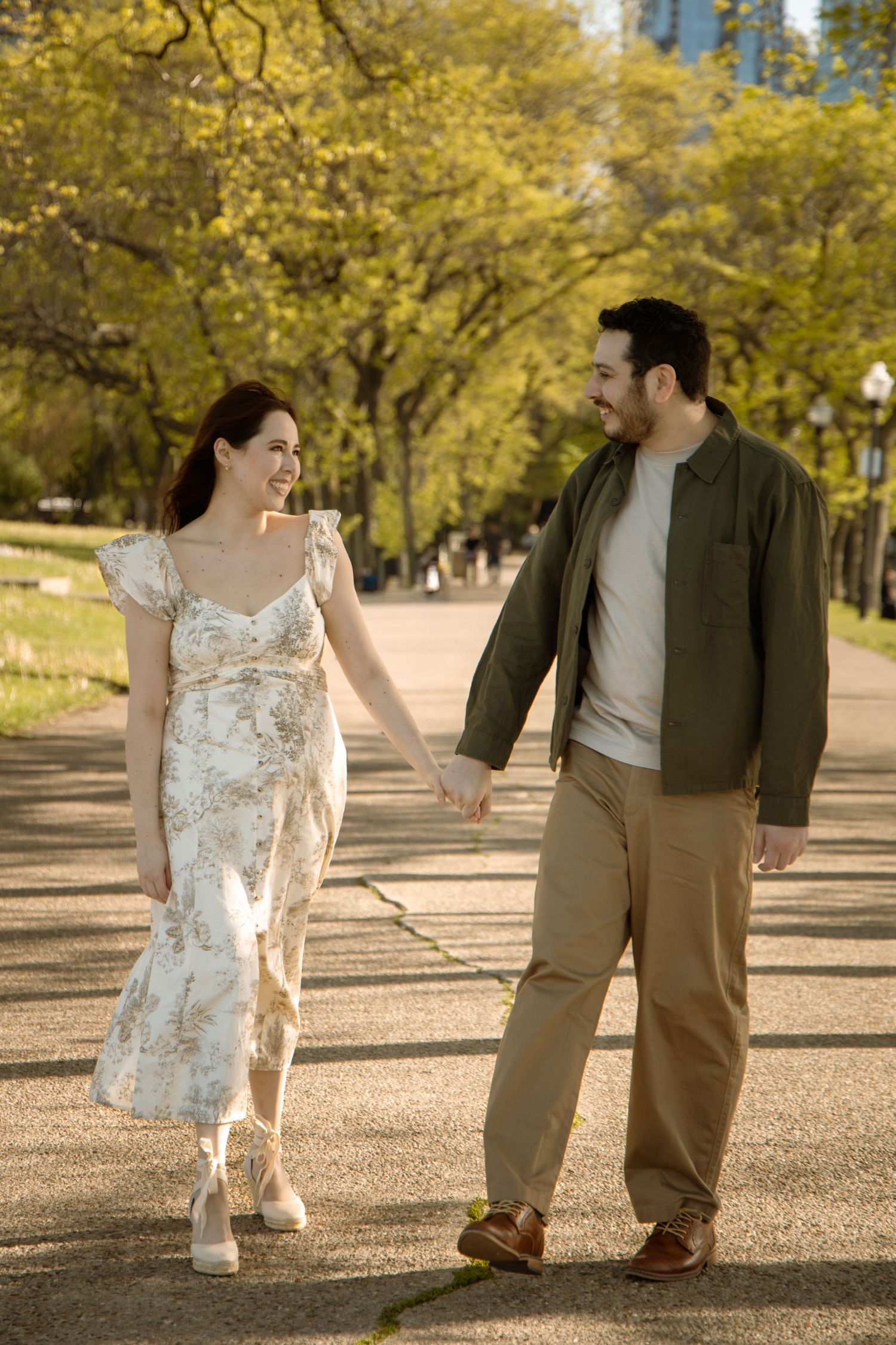 Couple holding hands and smiling while walking along a tree-lined park path on a sunny day.