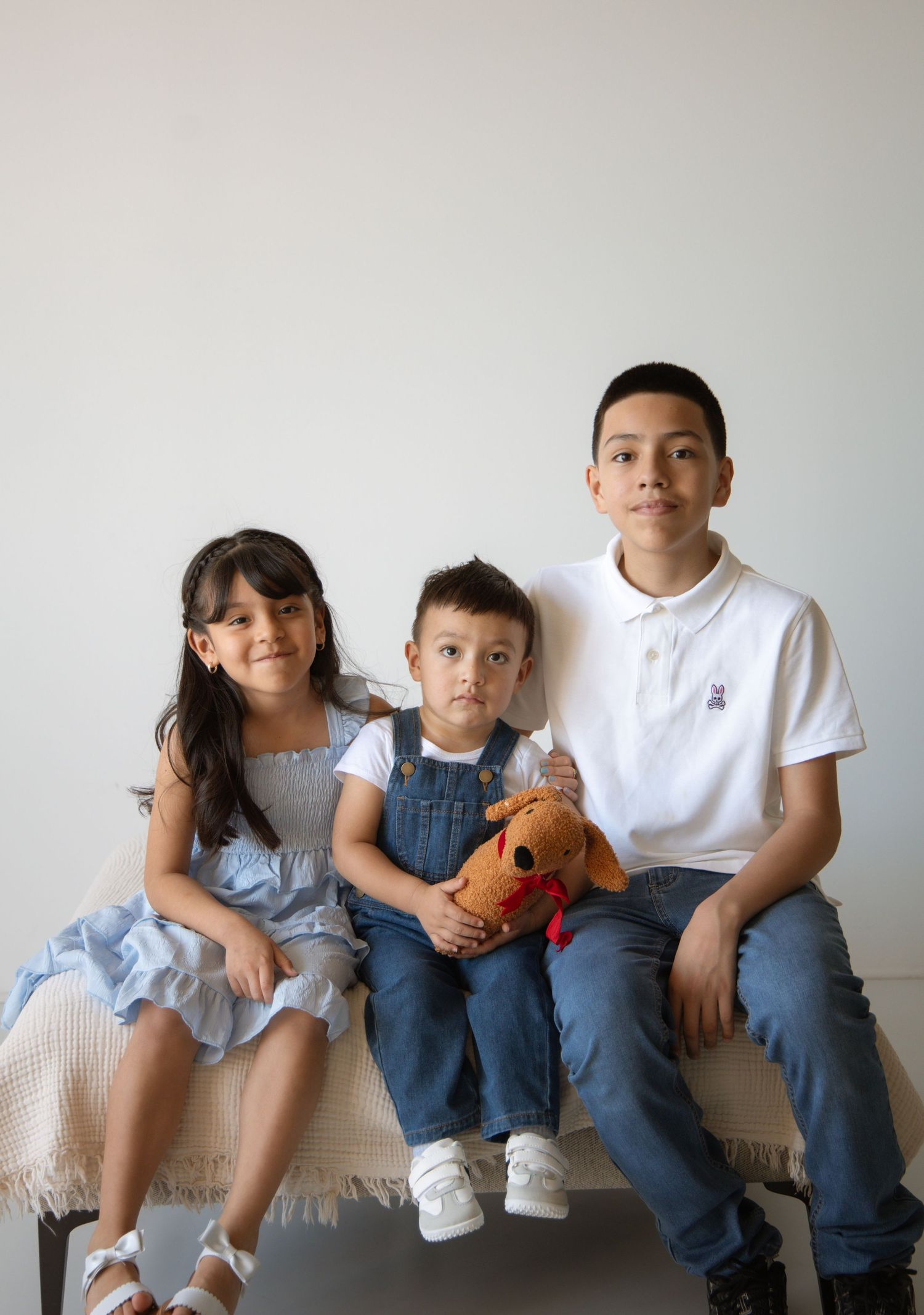 Three children sitting together on a bench, two older kids flanking a toddler holding a stuffed dog toy.