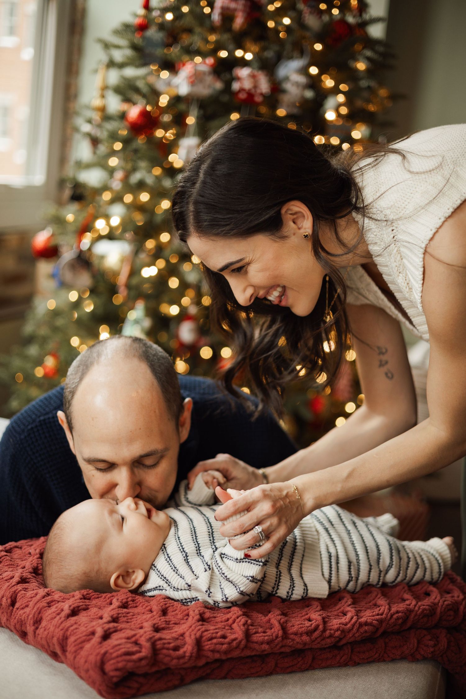 Happy parents with their baby by a decorated Christmas tree, father kissing infant's hand.