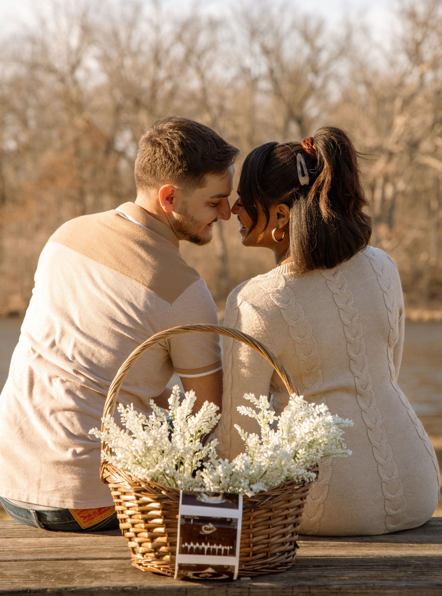Couple sitting back-to-back near a wicker basket of white flowers by a lake.