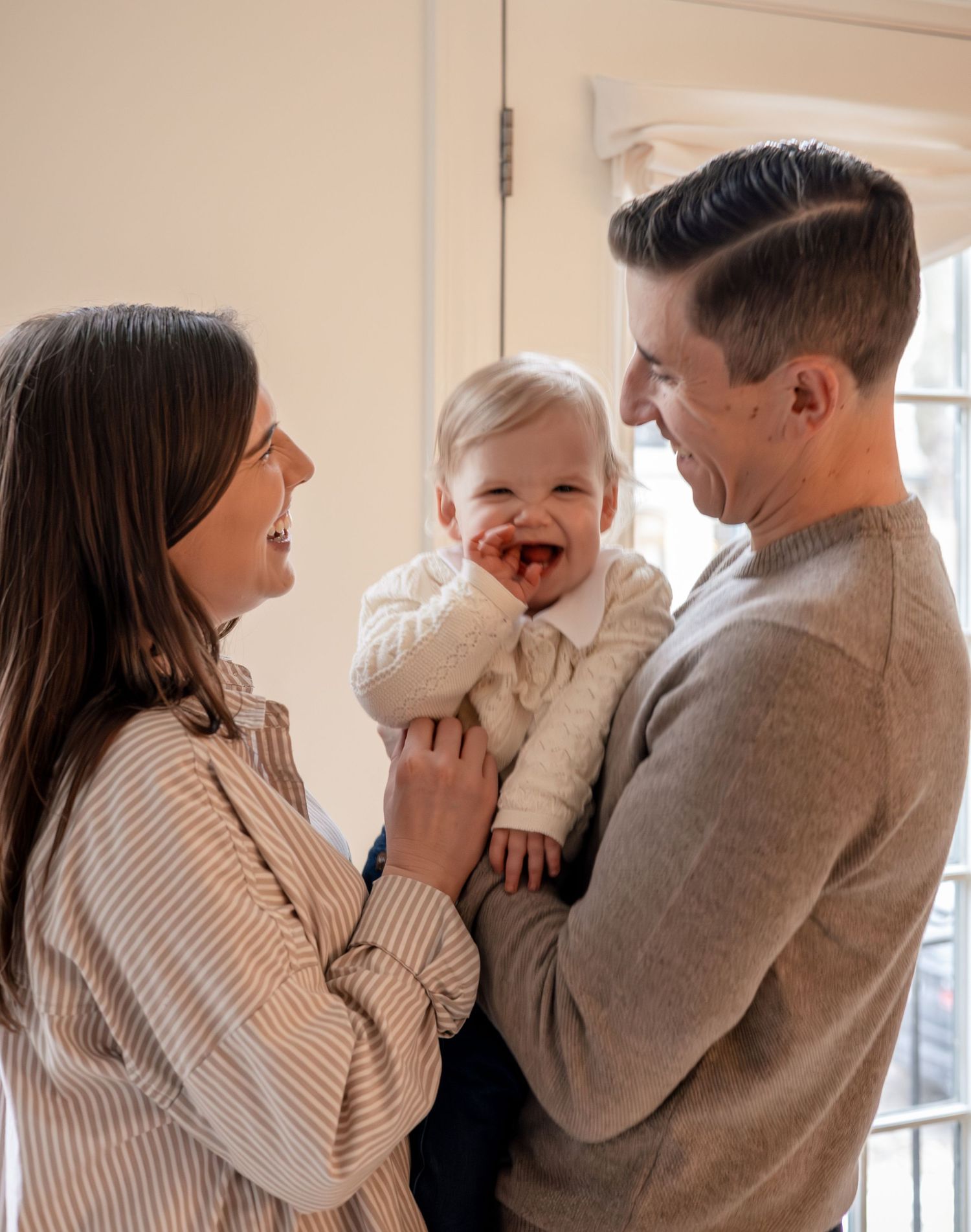 Laughing baby held by father while mother smiles in bright home interior.