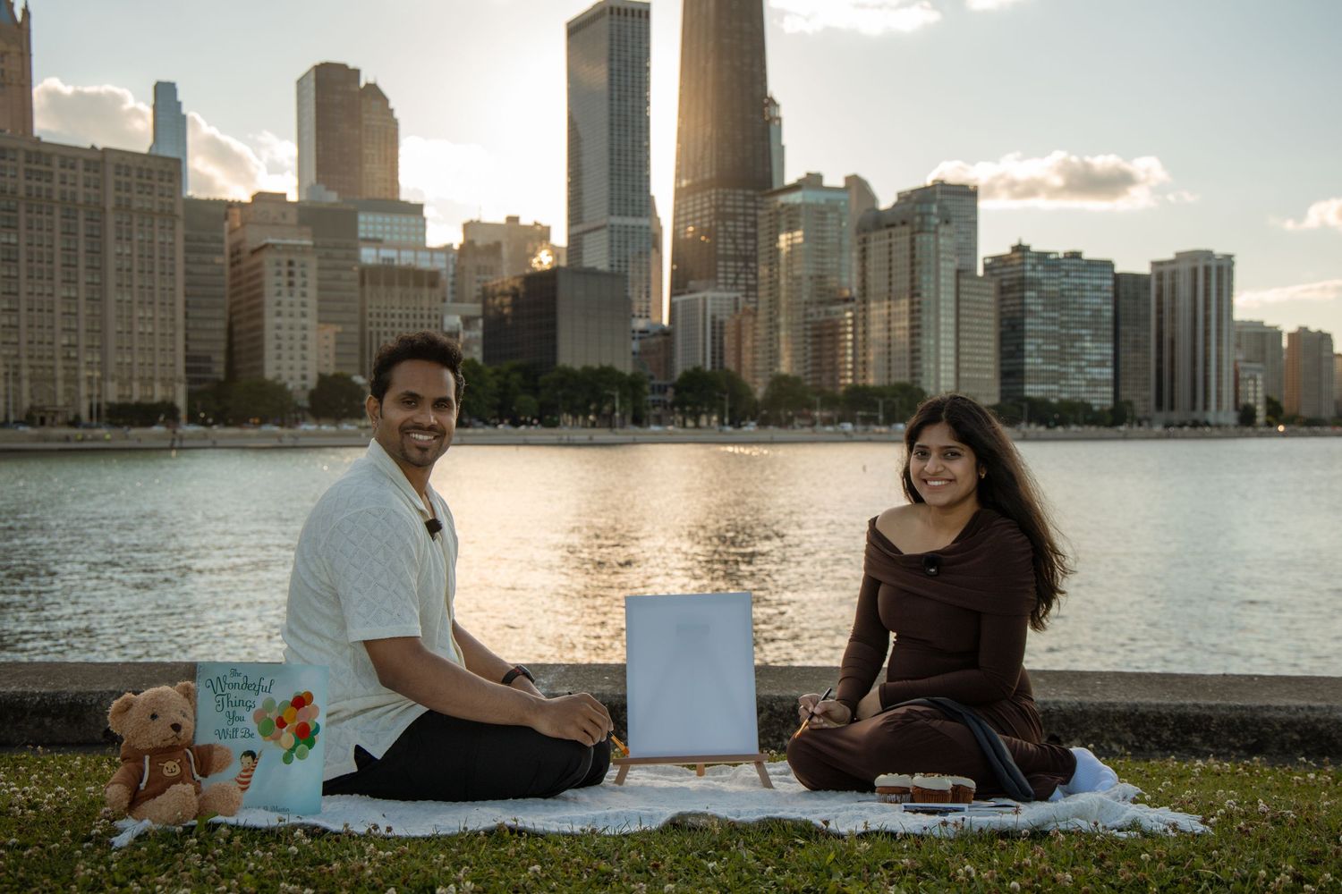 Couple enjoying a romantic picnic by the Chicago skyline waterfront at sunset with a teddy bear and gifts.