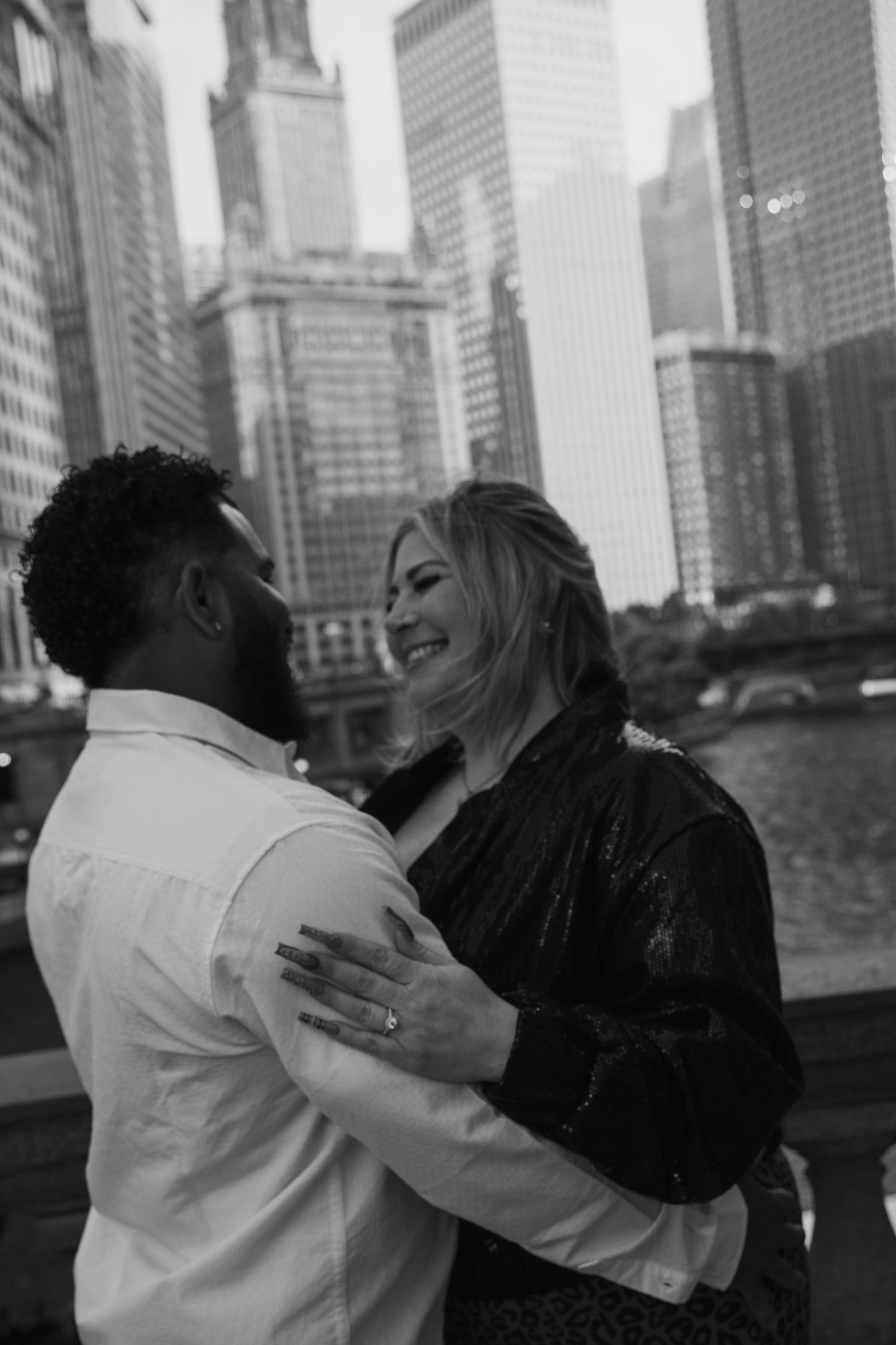 Black and white photo of a smiling couple embracing in front of Chicago city skyline and river.