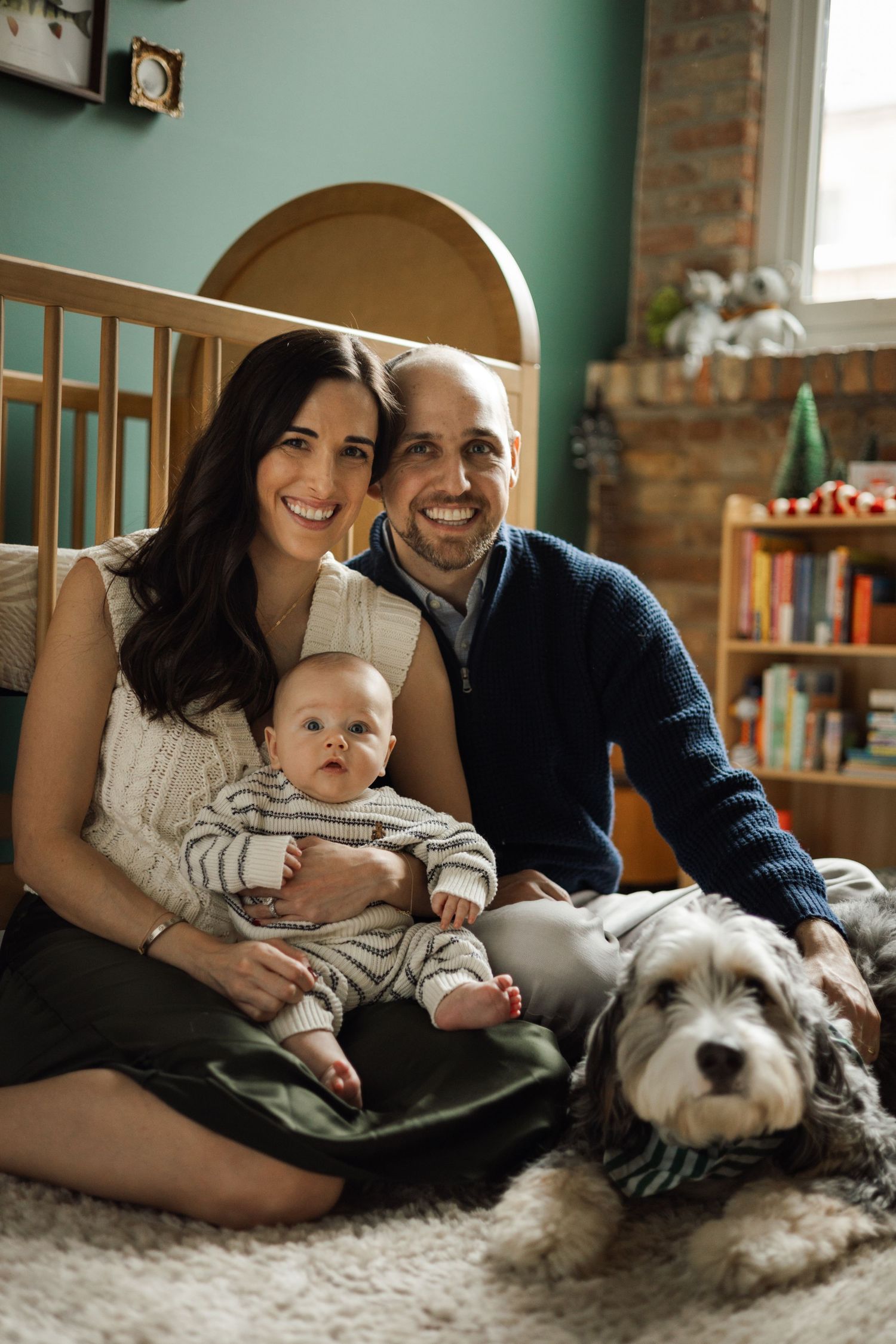 Happy family portrait with parents, baby, and fluffy dog sitting on floor in cozy nursery room.