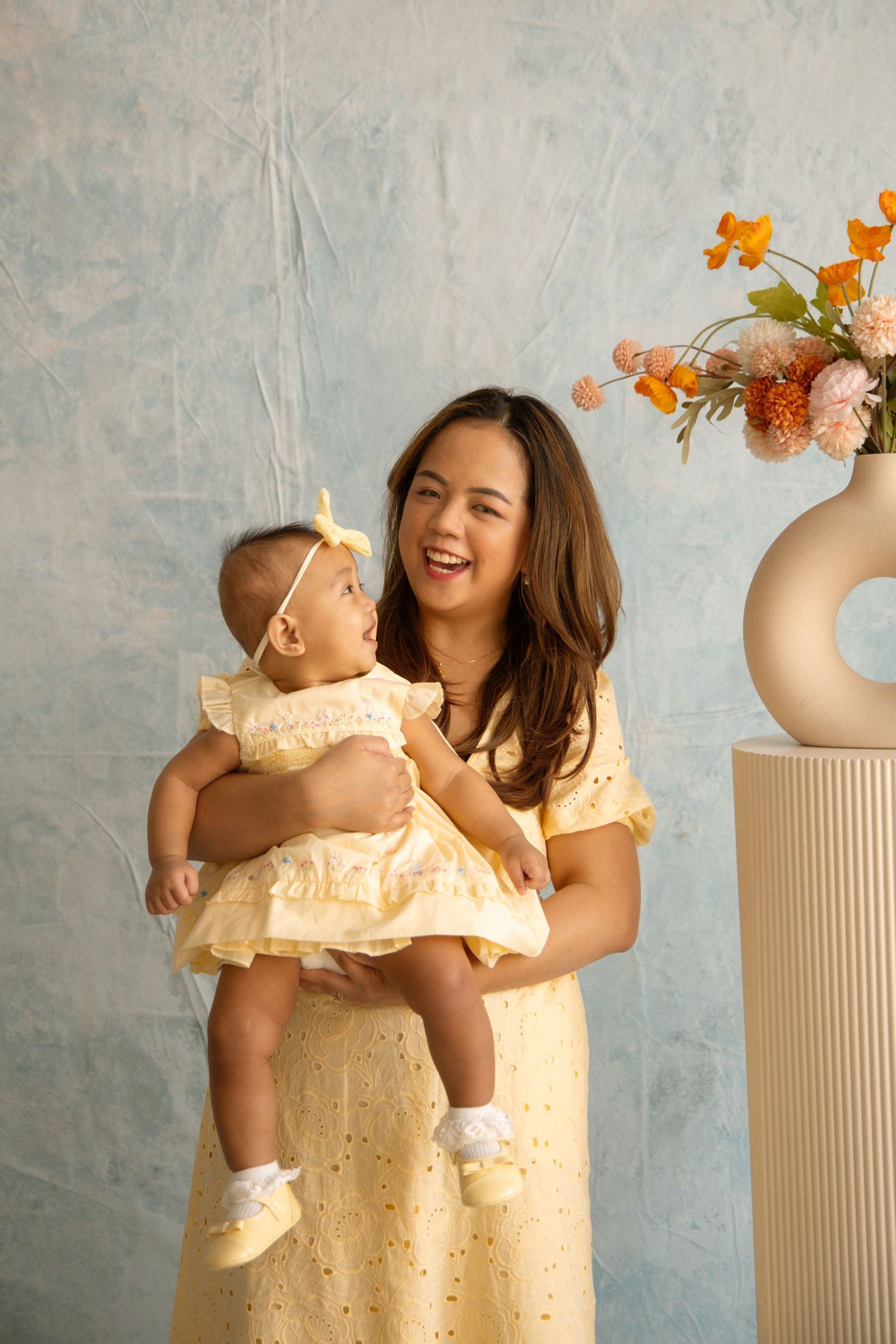 A smiling woman in a yellow dress holds a baby girl in a matching outfit near a floral arrangement.