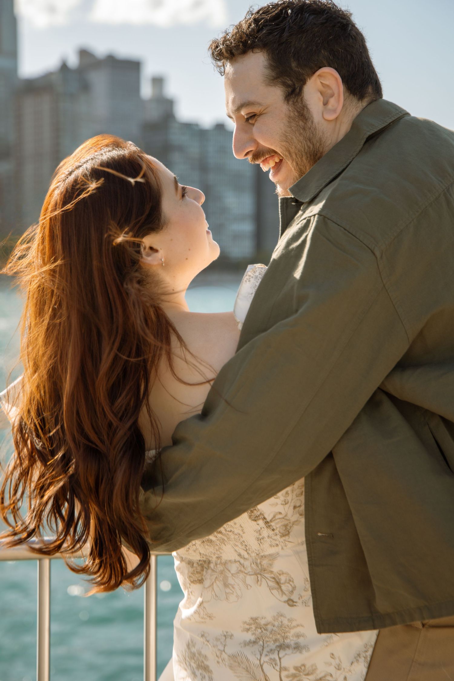 A couple embraces and smiles at each other on a waterfront, woman in floral dress, man in olive jacket.