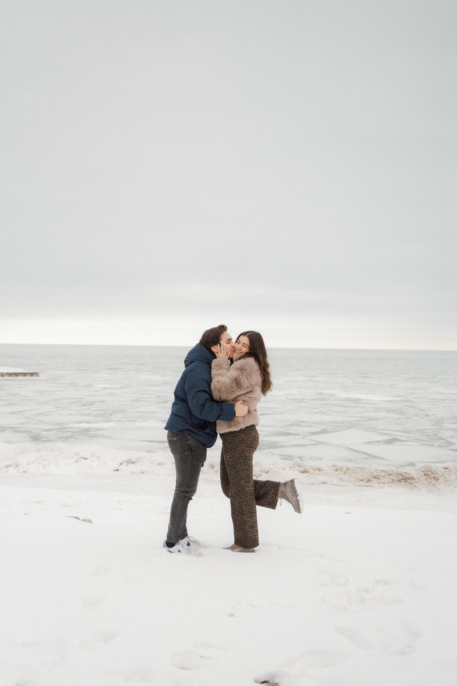 Couple shares a romantic kiss on a snowy lakefront beach in winter.