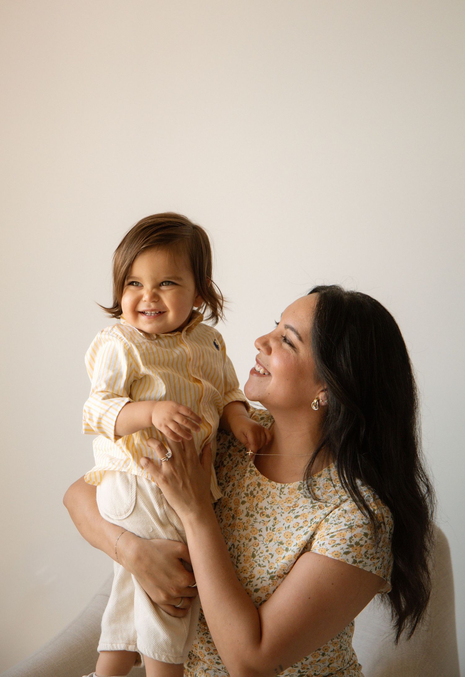 A woman in a floral top smiles while holding a happy baby in a yellow outfit against a white background.